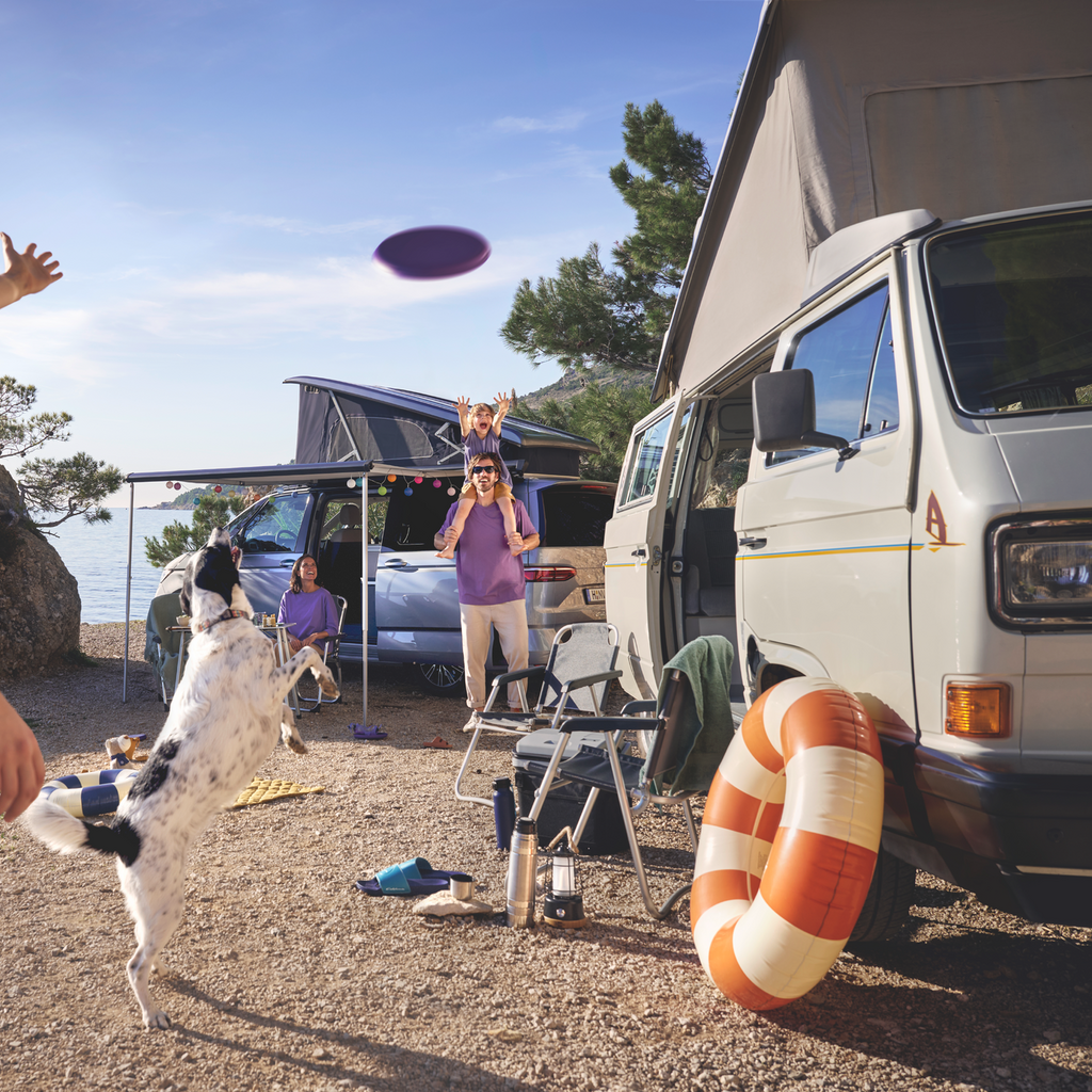 Eine Familie spielt auf einem Campingplatz, im Hintergrund ist ein Volkswagen California zu sehen.