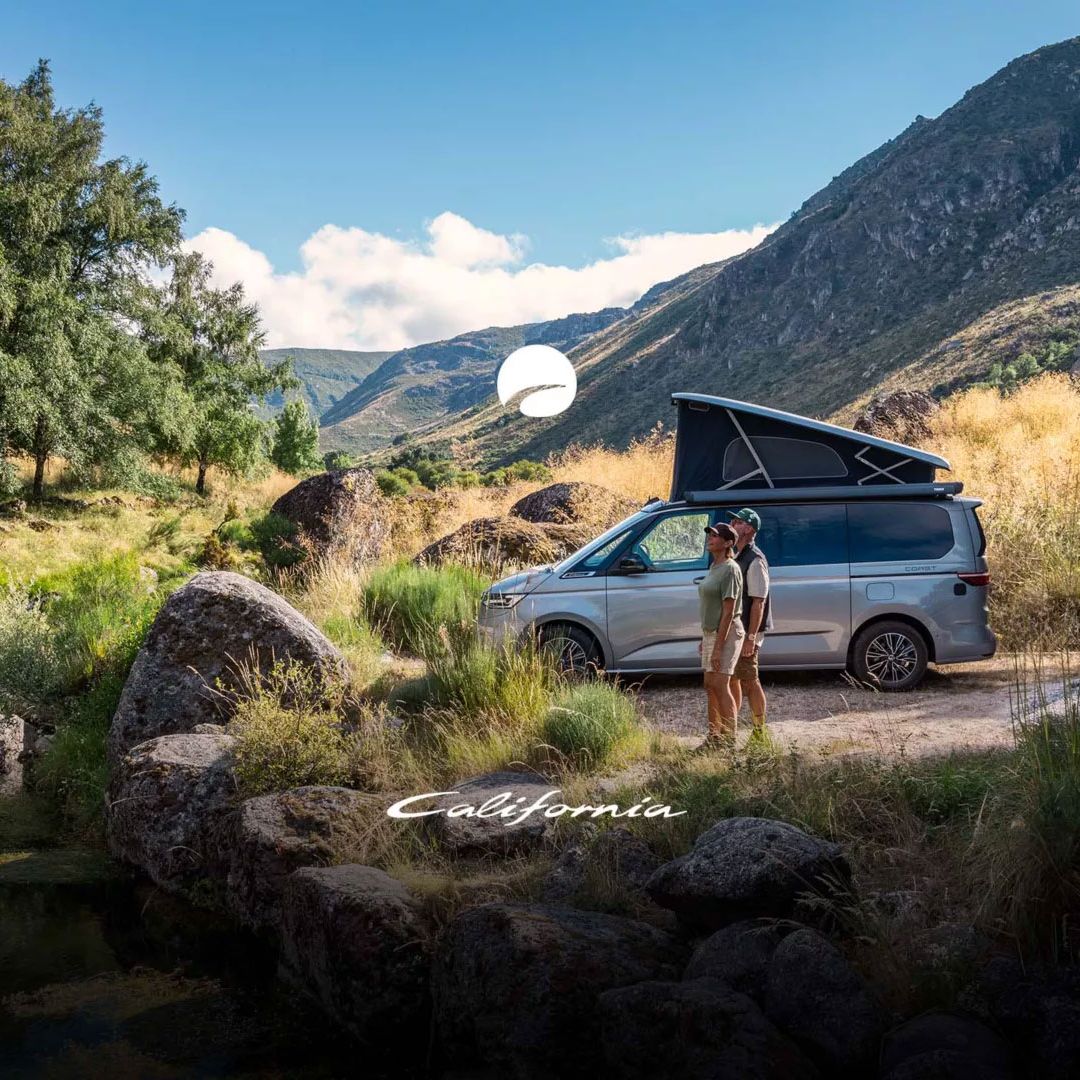 Ein Paar steht neben einem VW California mit ausgeklapptem Dach in einer malerischen Landschaft. Es gibt Berge im Hintergrund und einen kleinen See auf der linken Seite. 
