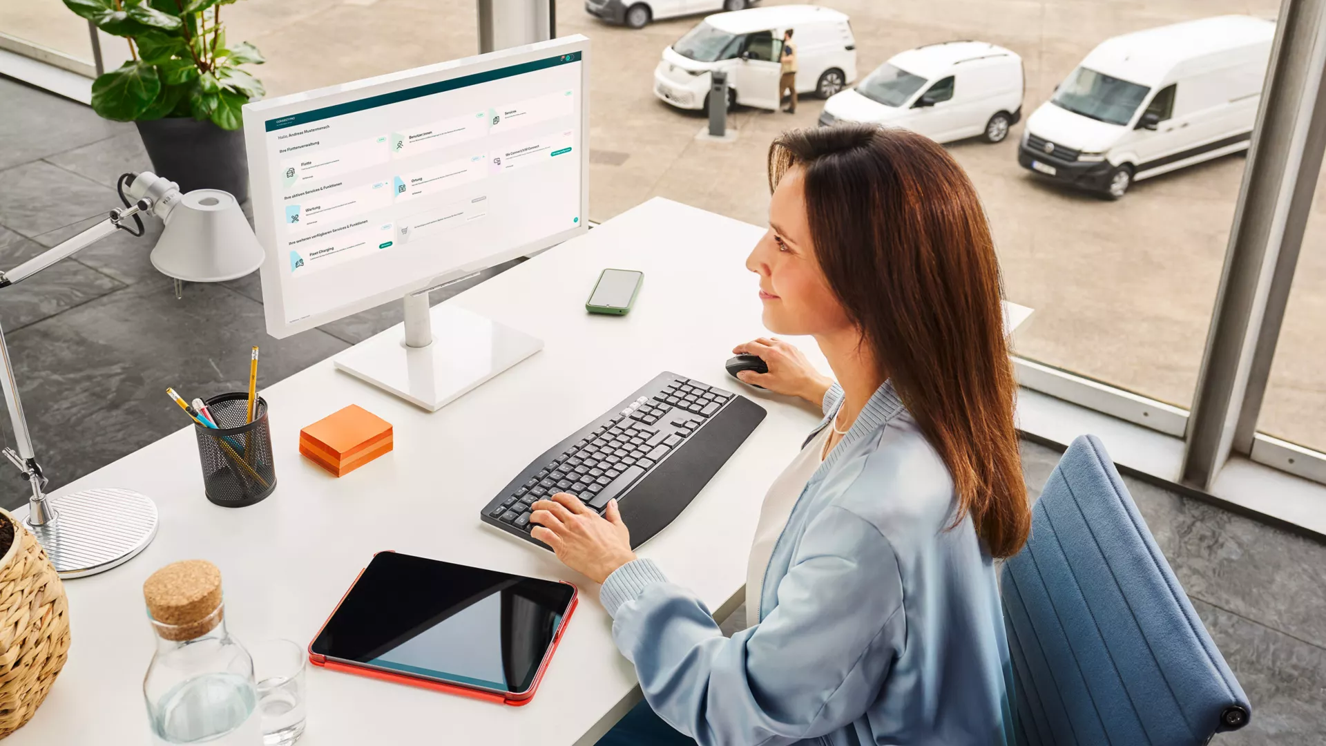 Eine Frau sitzt an einem Schreibtisch vor einem Computer in einem modernen Büro mit großen Fenstern. Draußen sind mehrere weiße Lieferwagen zu sehen. Auf dem Schreibtisch liegen eine Wasserflasche, ein Tablet und Schreibutensilien.