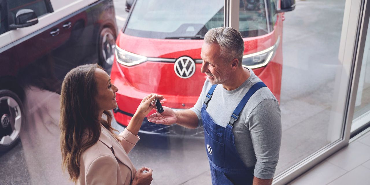 Ein Volkswagen Techniker überreicht einer Kundin ihren Autoschlüssel. Im Hintergrund befindet sich ein roter Volkswagen ID. Buzz.