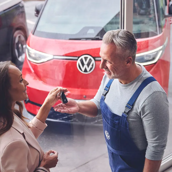Ein Volkswagen Techniker überreicht einer Kundin ihren Autoschlüssel. Im Hintergrund befindet sich ein roter Volkswagen ID. Buzz.