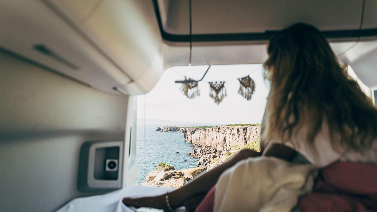 Eine Person sitzt in einem Volkswagen Grand California am Rand eines Bettes und blickt durch die offene Heckklappe auf eine felsige Küste.