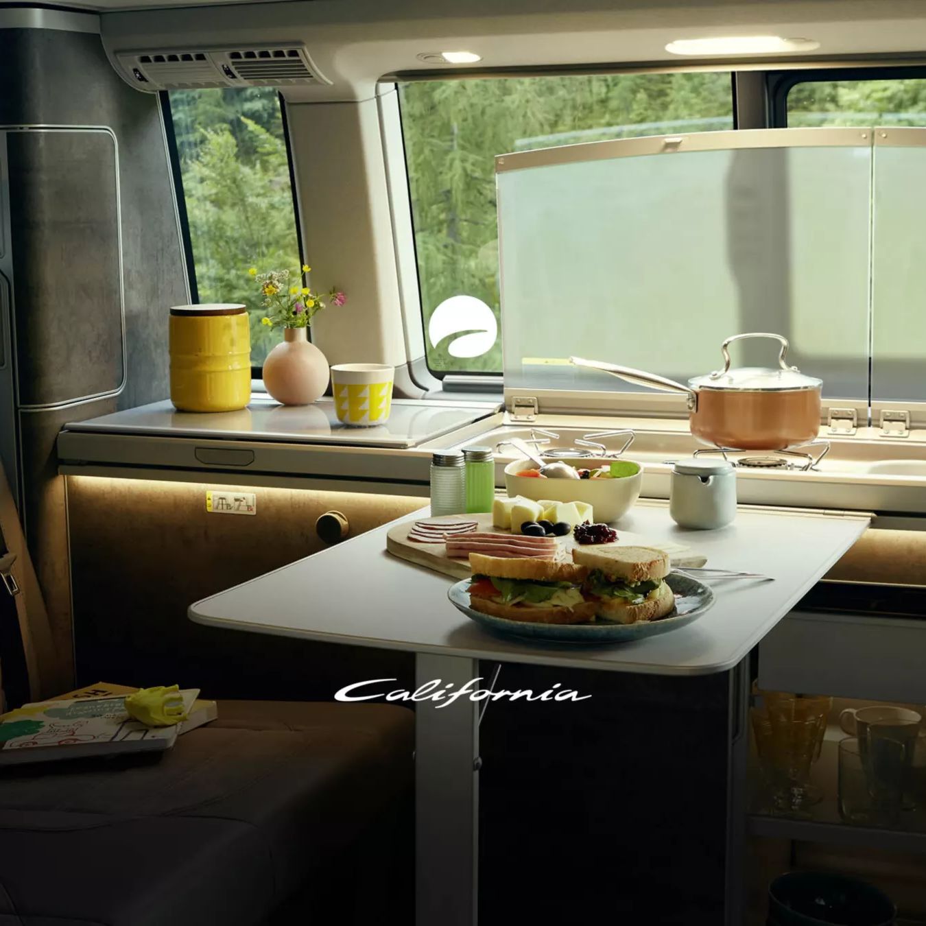 Das Bild zeigt das Innere eines VW California mit einem kleinen Tisch, auf dem Frühstück serviert wird. Im Hintergrund sieht man das Spülbecken sowie sonstige Küchenutensilien. Durch das Fenster ist viel Grün zu erkennen.