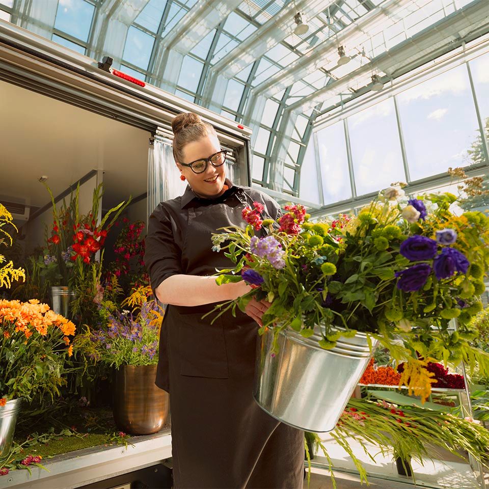 Eine Person steht in einem Blumenladen, umgeben von einer Vielzahl von bunten Blumenarrangements. Sie hält einen silbernen Eimer voller frischer Blumen. Der Raum hat große Fenster, die viel Tageslicht hereinlassen.