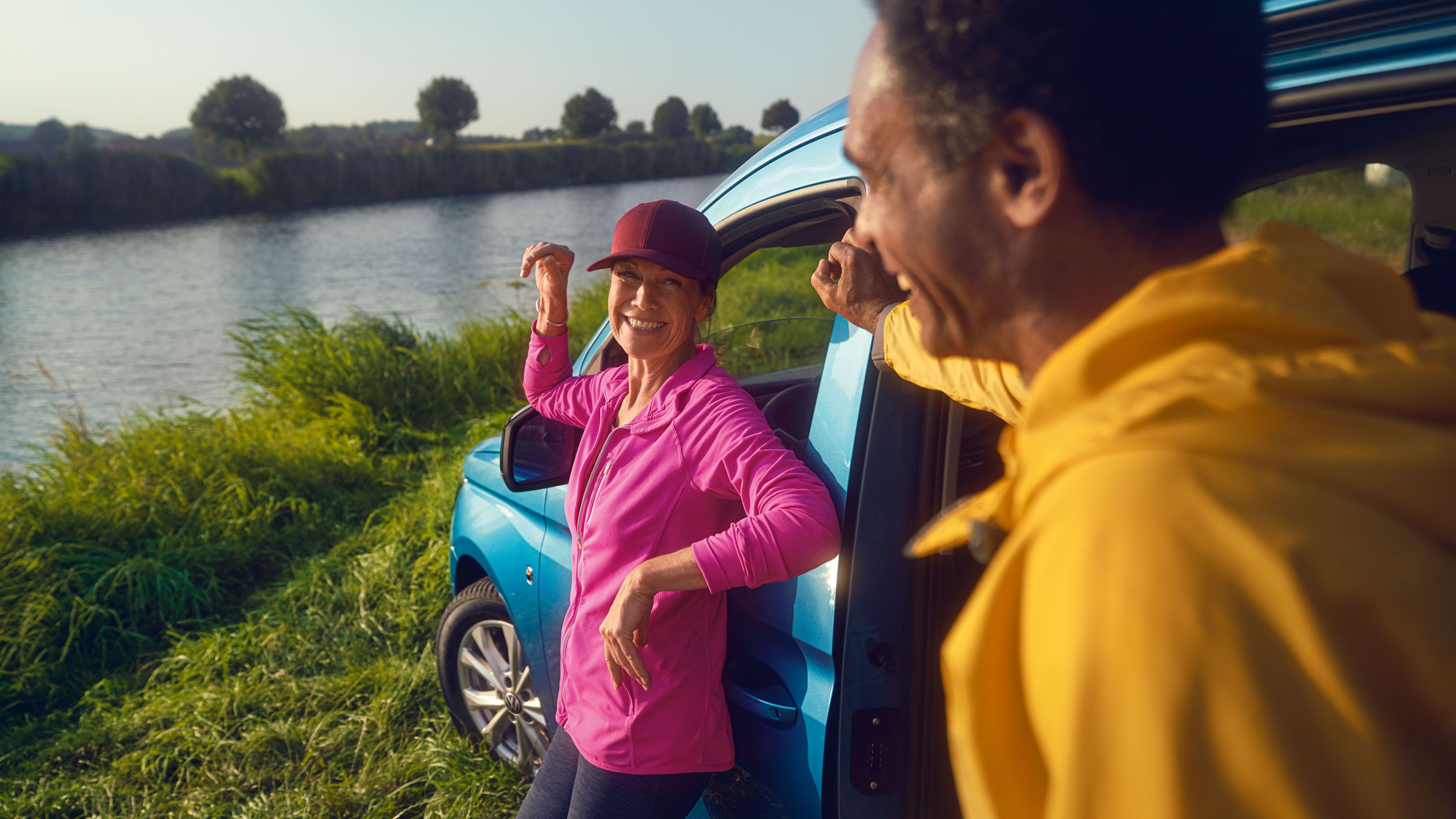 Mann und Frau lehnen an Seite eines blauen VW Caddys, der an einem See steht