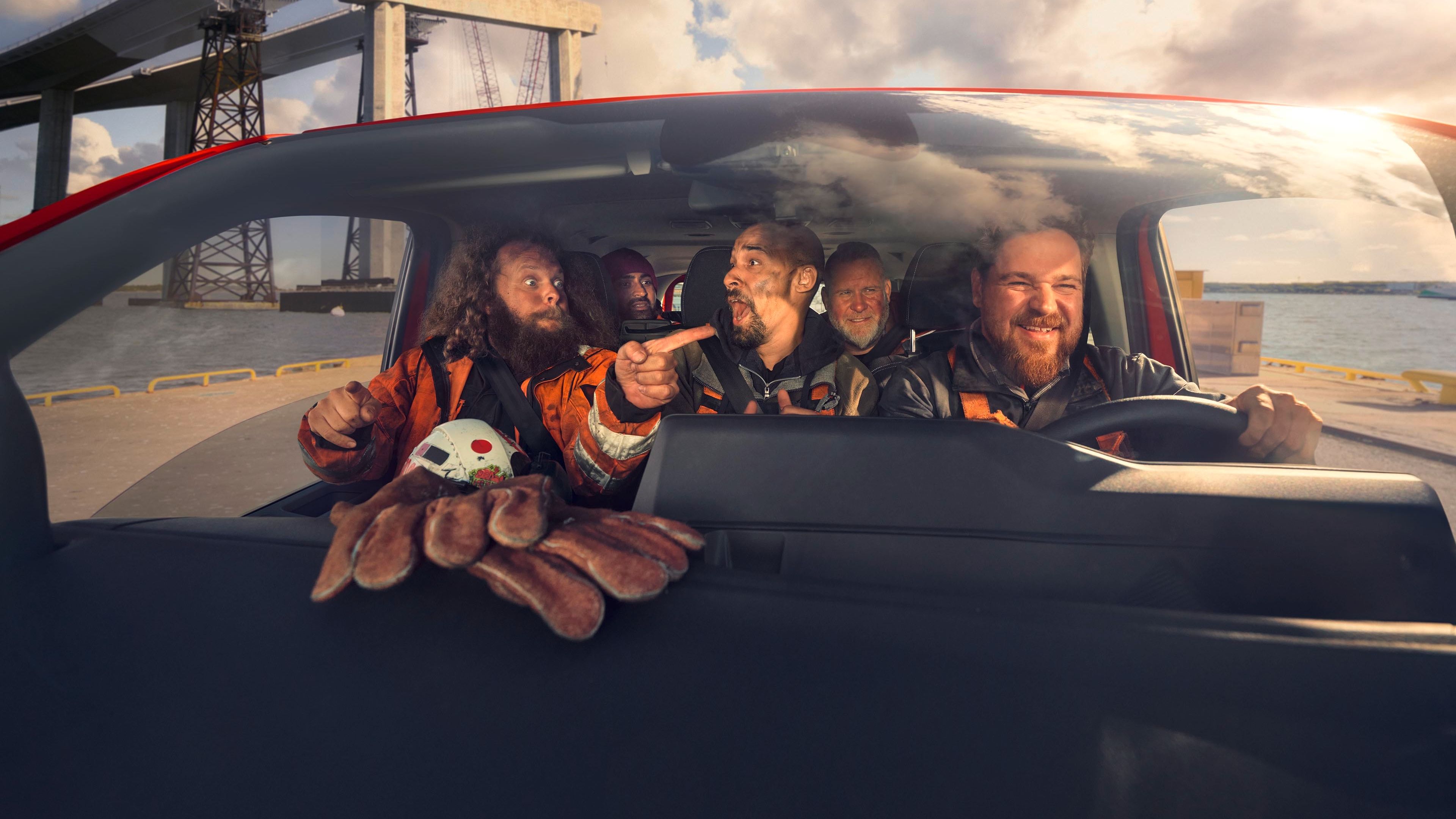 Mehrere Personen in Sicherheitskleidung sitzen in einem Auto und scheinen sich eine Baustelle mit Kranen und Stahlkonstruktionen anzusehen. Eine Person zeigt mit dem Finger in Richtung der Baustelle. Auf der Armaturenbrettablage liegt eine Spielzeugkrabbe.