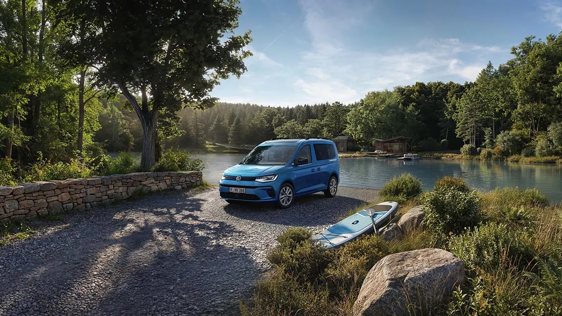 Ein blauer Volkswagen Caddy steht auf einem Schotterweg am Ufer eines ruhigen Sees, umgeben von dichtem, grünem Wald.