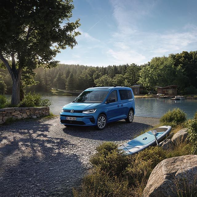 Ein blauer Volkswagen Caddy steht auf einem Schotterweg am Ufer eines ruhigen Sees, umgeben von dichtem, grünem Wald.