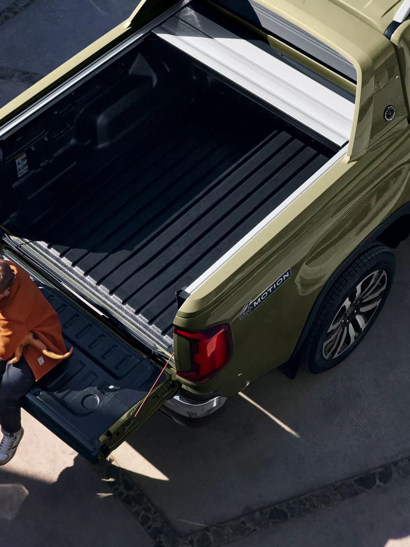 Zwei Kinder sitzen auf der heruntergeklappten Ladefläche eines olivgrünen Pickup-Trucks und benutzen ein Tablet. Die Ladefläche des Fahrzeugs ist leer und mit einer schwarzen Matte ausgekleidet. Die Szene findet draußen statt, bei natürlichem Licht.