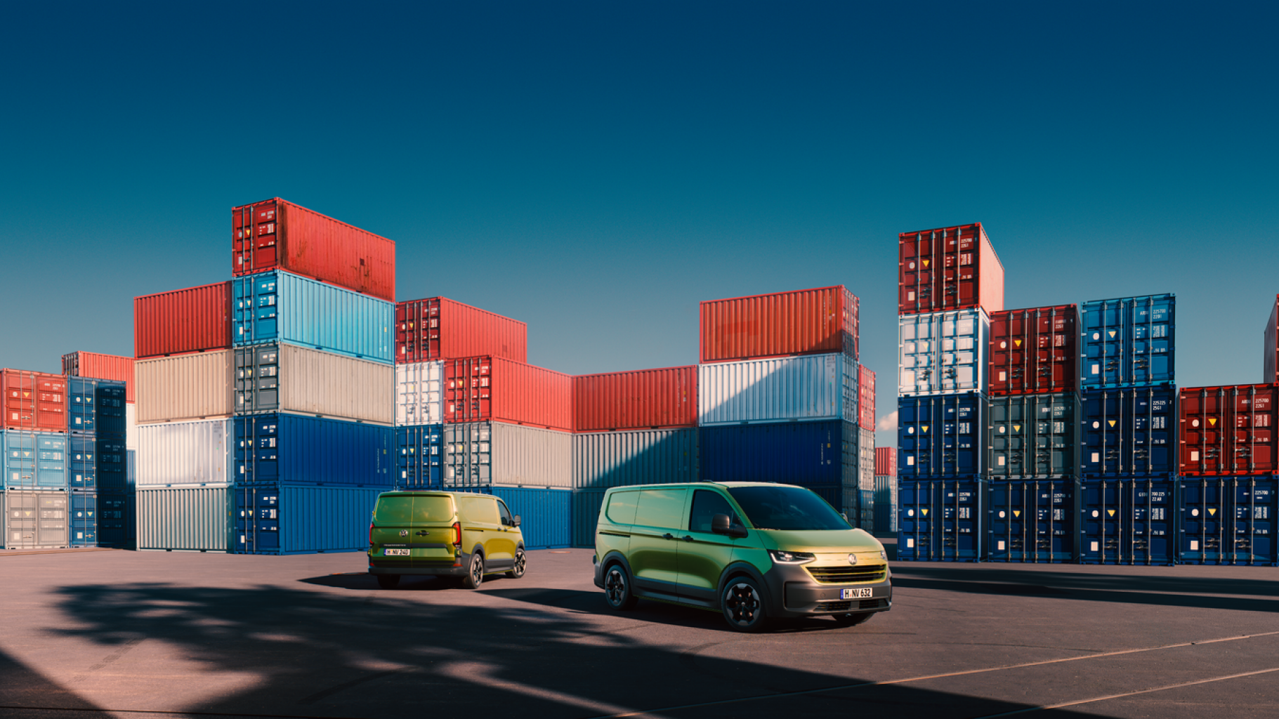 Zwei grüne Volkswagen Transporter vor gestapelten Containern in einem Hafen.