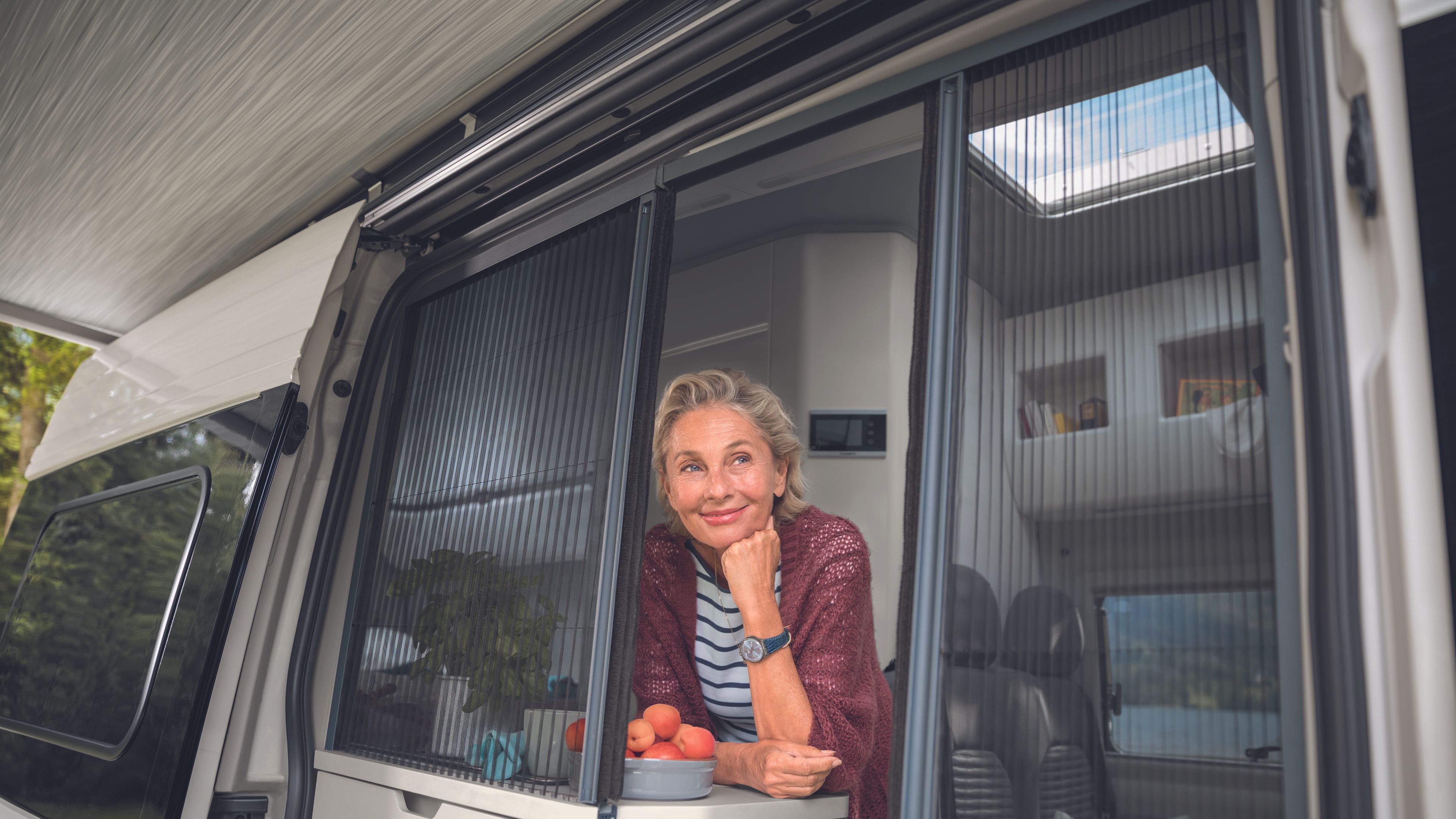 Eine Frau stützt sich auf dem Küchenschrank ab und blickt durch die offene Schiebetür. Rechts und links befinden sich Moskitonetze. 