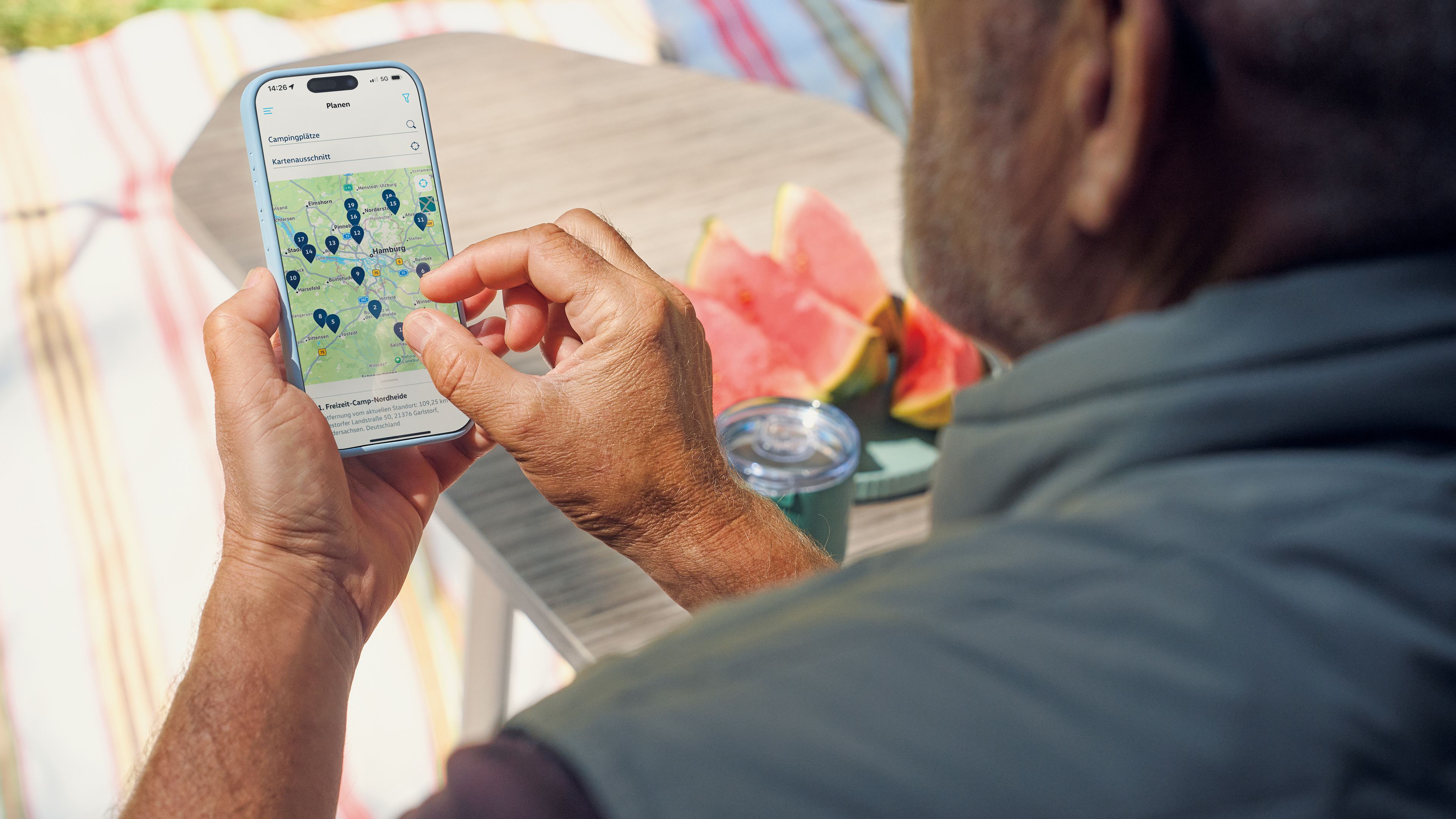 Ein älterer Mann nutzt ein Smartphone, um eine Karte mit mehreren Markierungen darauf anzusehen. Im Hintergrund sieht man einen Tisch mit Wassermelonenstücken und einen Streifenteppich.