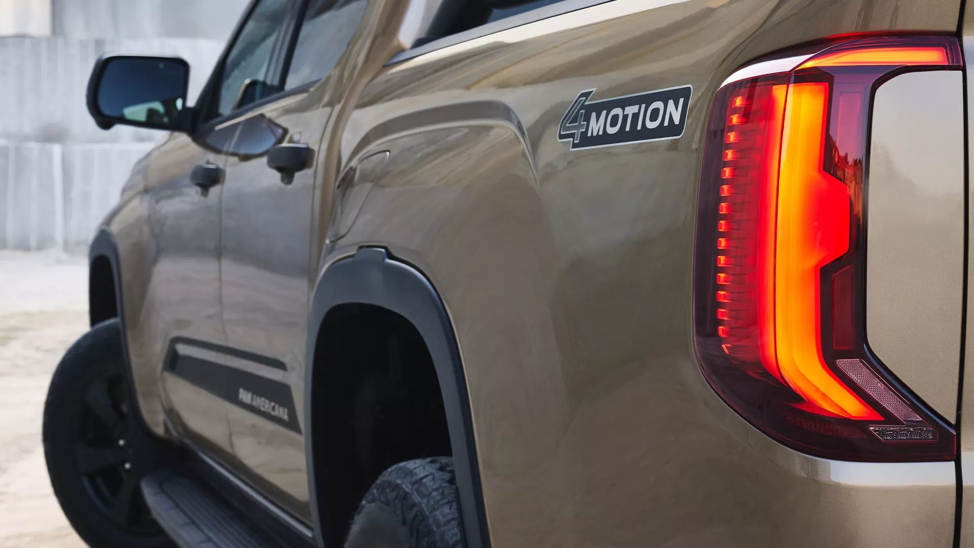 Ein brauner Pickup-Truck ist seitlich und von hinten zu sehen. Auf der Karosserie befinden sich die Schriftzüge "4MOTION" und "Panamericana". Das Rücklicht des Fahrzeugs ist beleuchtet.