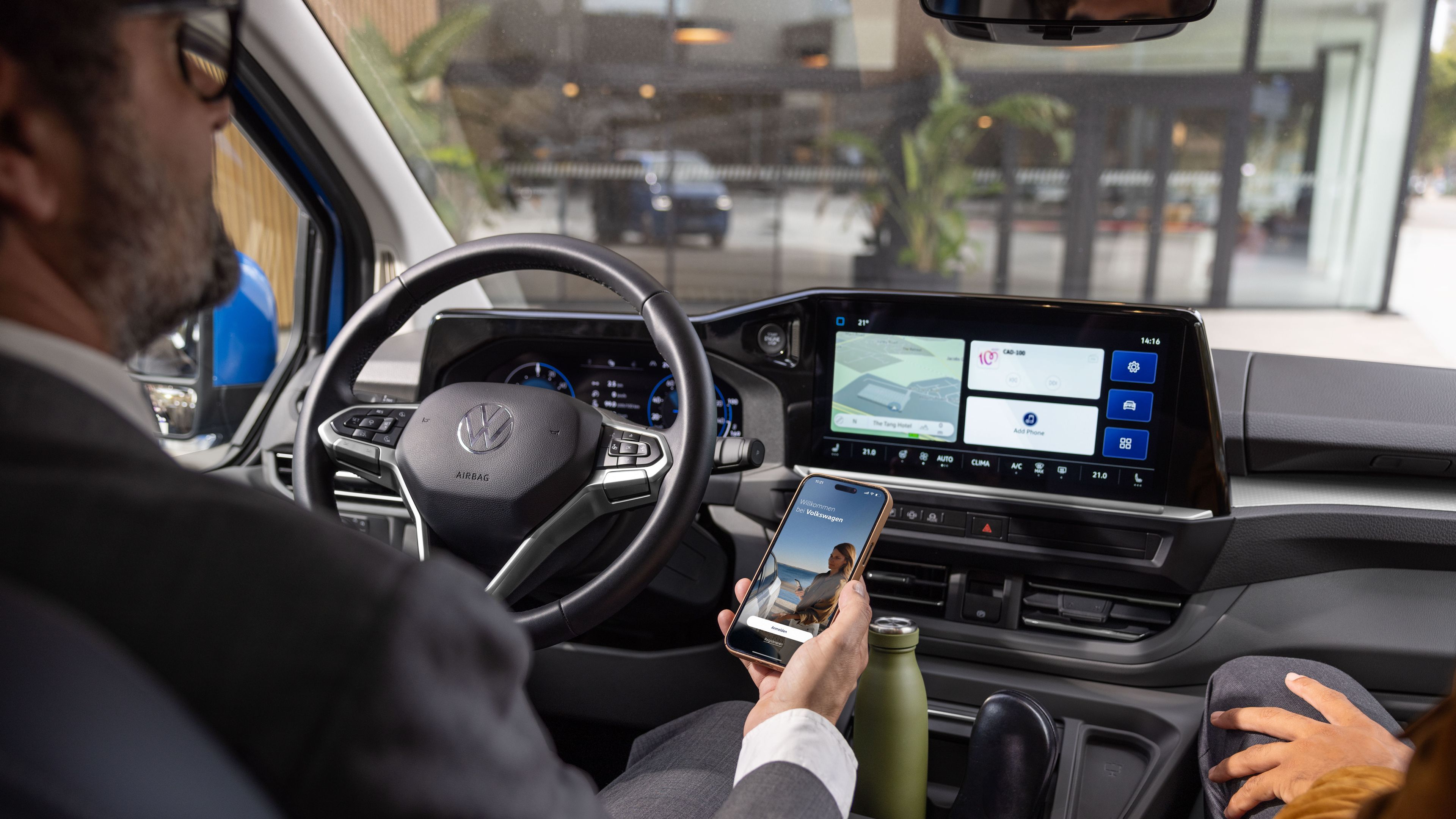 Ein Mann sitzt am Steuer eines Volkswagen und hält ein Smartphone in der Hand. Das Armaturenbrett zeigt ein großes Display mit verschiedenen Navigations- und App-Optionen. Eine weitere Person sitzt auf dem Beifahrersitz.