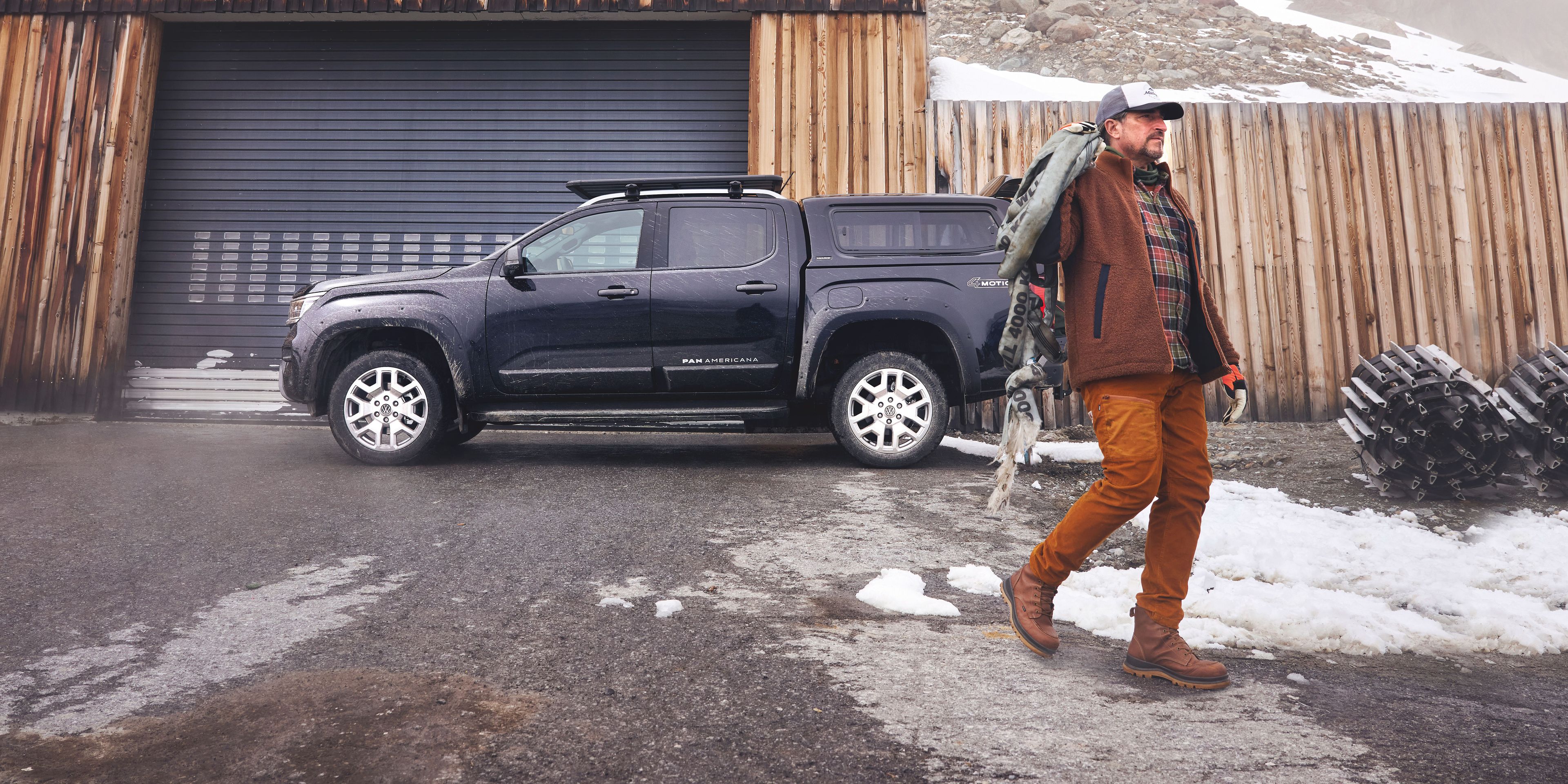 Ein Mann in Outdoor-Kleidung geht auf einem nassen, teils verschneiten Platz. Hinter ihm steht ein dunkler Volkswagen Amarok vor einer hölzernen Garage, im Hintergrund ragen nebelverhangene Felsen auf.