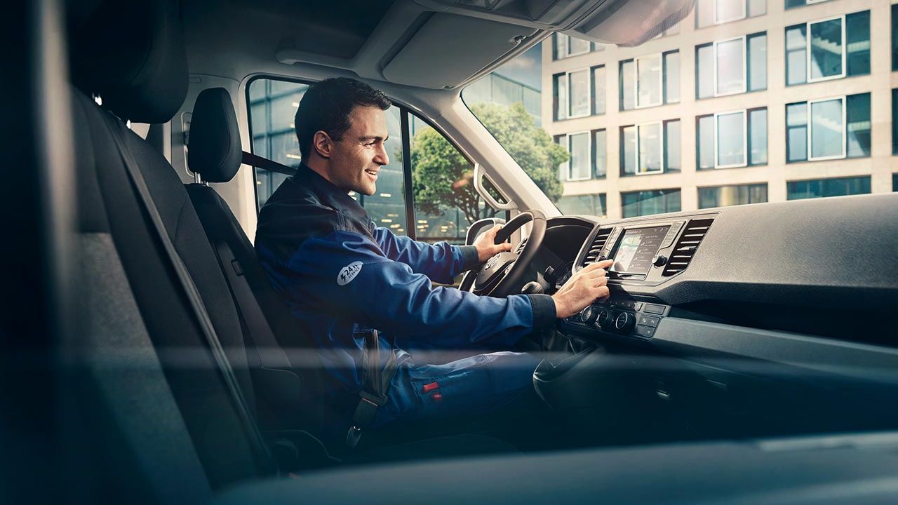 Eine Person sitzt am Steuer eines Volkswagen Nutzfahrzeugs und trägt eine blaue Arbeitsuniform. Im Hintergrund sieht man ein modernes Bürogebäude und Bäume.