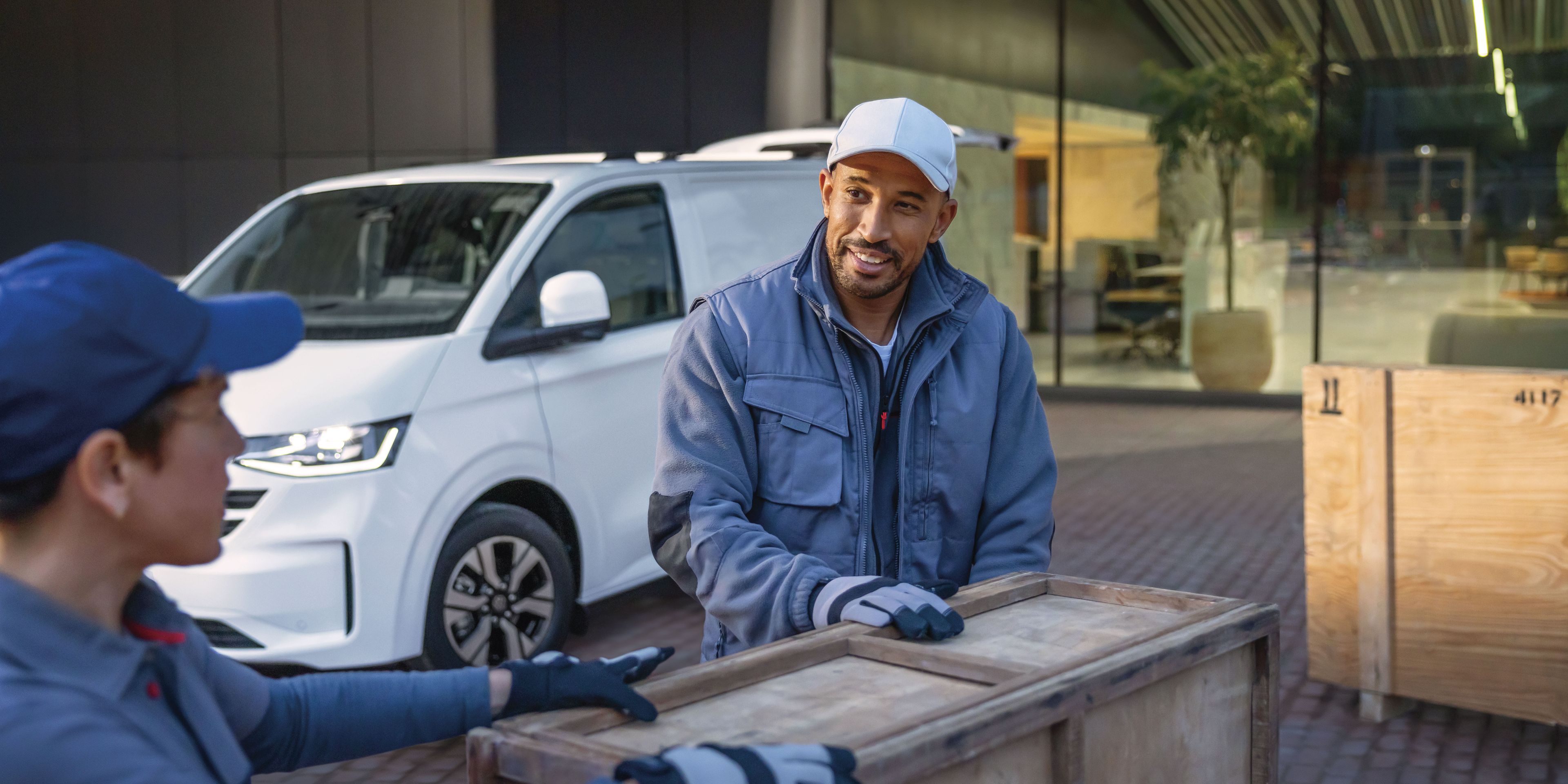 Zwei Männer tragen eine Holzkiste. Im Hintergrund ist ein weißer Volkswagen Transporter zu sehen.