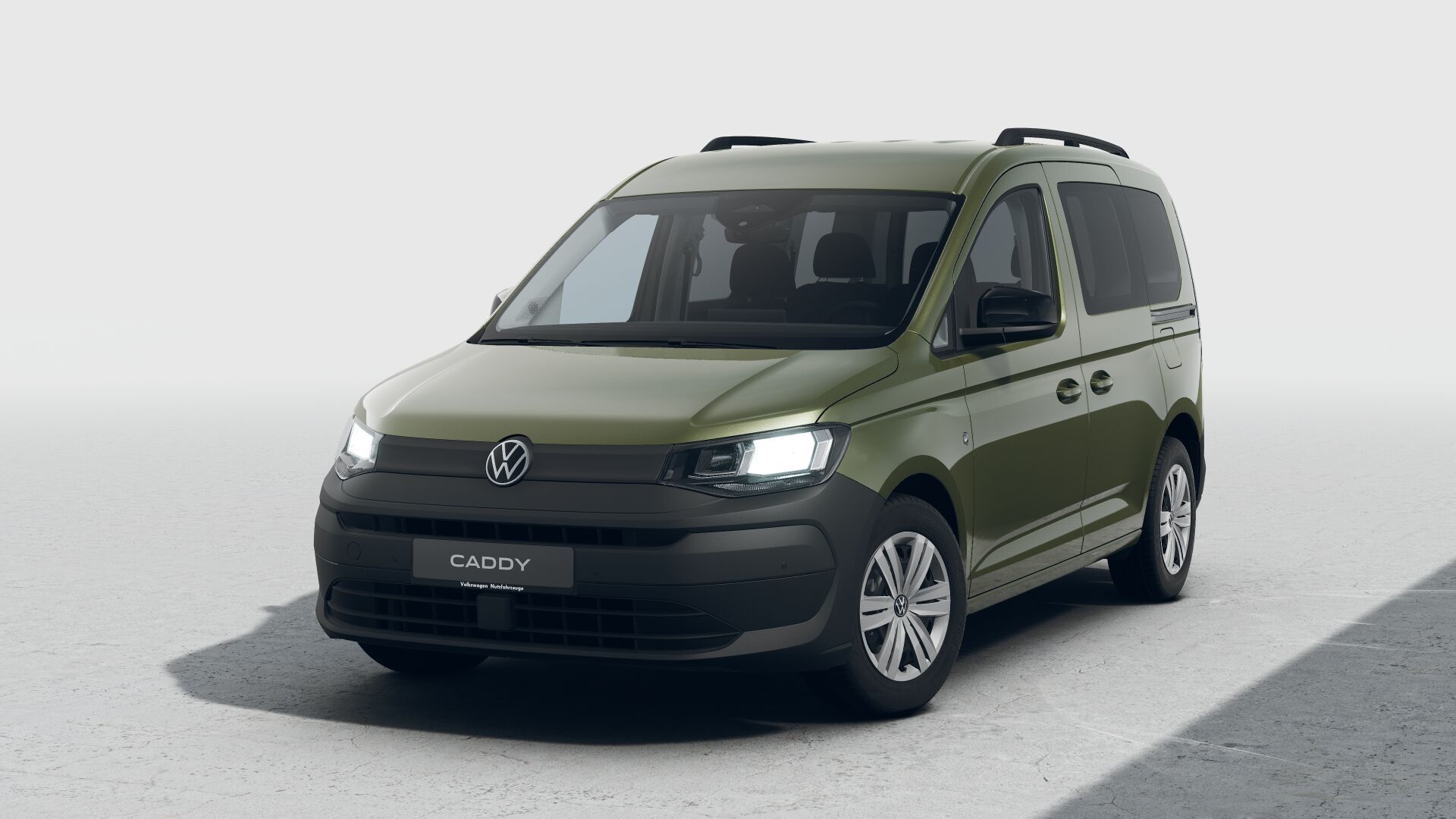 Ein olivgrüner VW Caddy steht schräg von vorn fotografiert auf einer hellen, neutralen Fläche. Das Fahrzeug hat fünf Türen, Dachreling und moderne, schmale Scheinwerfer. Auf dem Kennzeichenbereich vorne steht der Modellname „CADDY“.