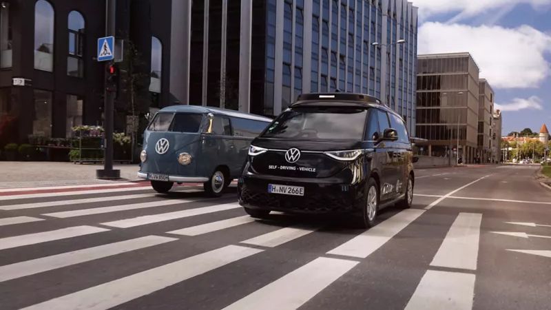 Ein moderner schwarzer VW Minivan und ein klassischer blauer VW Bus fahren auf einer Straße nebeneinander. Die Straße führt durch ein Geschäftsviertel mit hohen Gebäuden. Beide Fahrzeuge überqueren einen Zebrastreifen.
