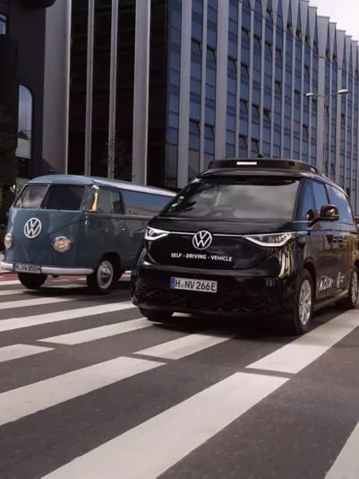 Ein moderner schwarzer VW Minivan und ein klassischer blauer VW Bus fahren auf einer Straße nebeneinander. Die Straße führt durch ein Geschäftsviertel mit hohen Gebäuden. Beide Fahrzeuge überqueren einen Zebrastreifen.