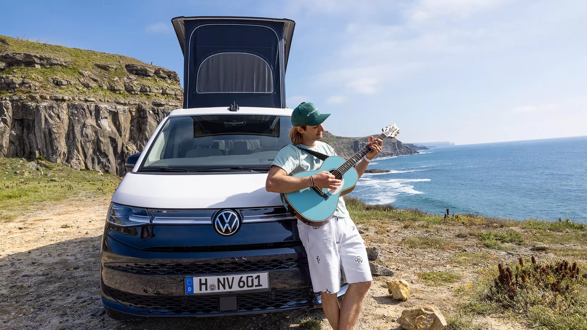 Ein Mann spielt Gitarre neben einem parkenden VW California an einer Küste mit Klippen und Meer im Hintergrund.