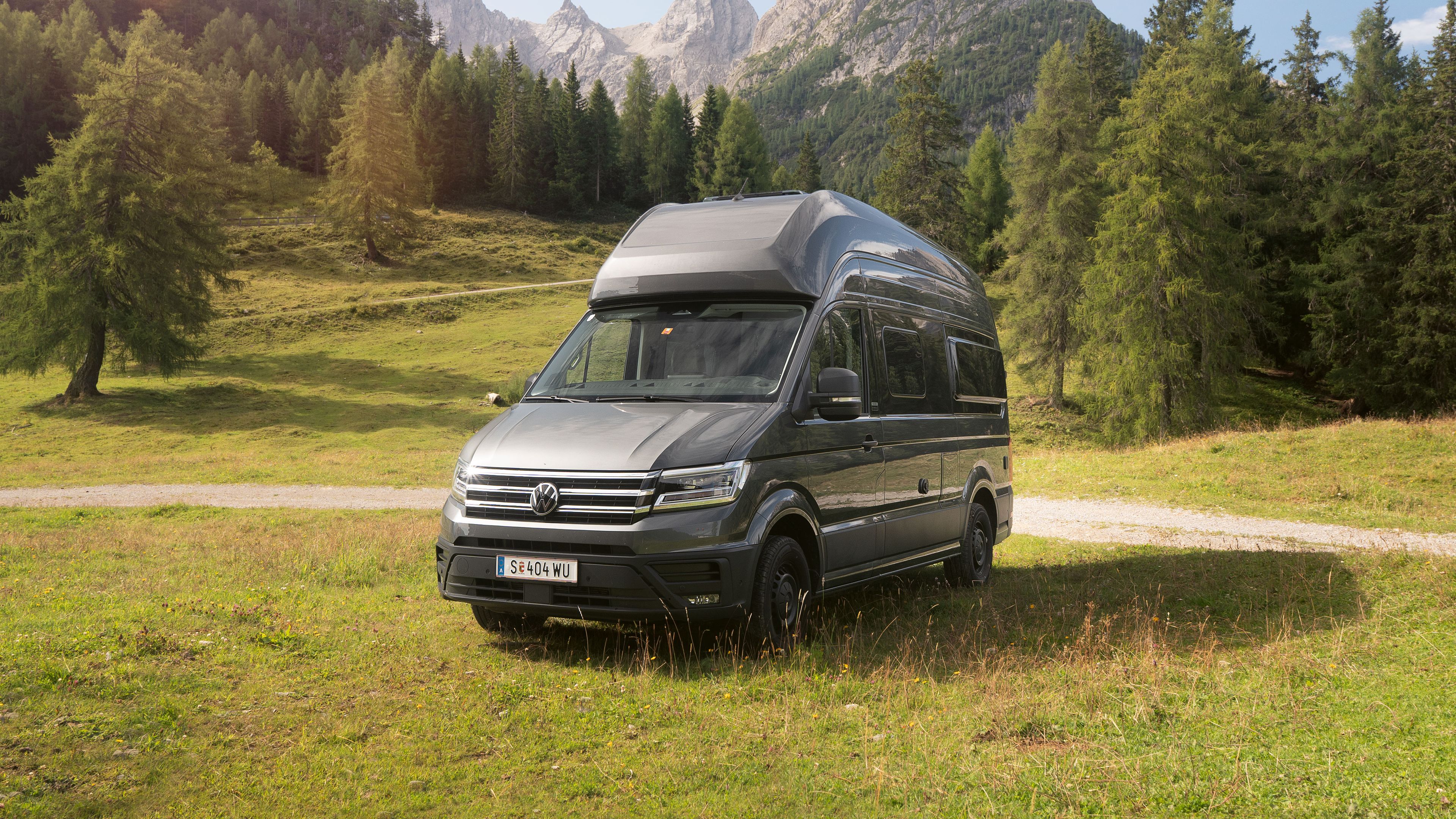 Ein silberner Wohnmobil-Van steht auf einer grünen Wiese vor einer malerischen Berglandschaft mit Bäumen und blauem Himmel.