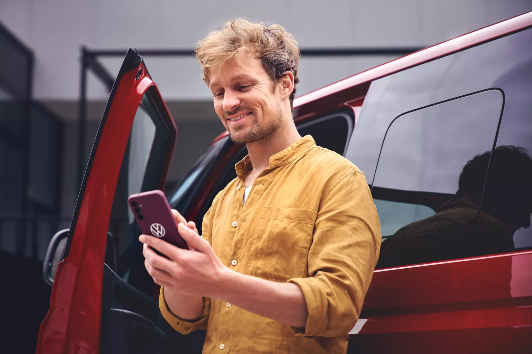 Ein Mann steht neben einem roten Auto und hält ein Smartphone in der Hand. Er trägt ein senfgelbes Hemd.