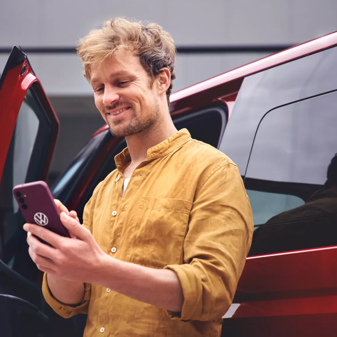 Ein Mann steht neben einem roten Auto und hält ein Smartphone in der Hand. Er trägt ein senfgelbes Hemd.