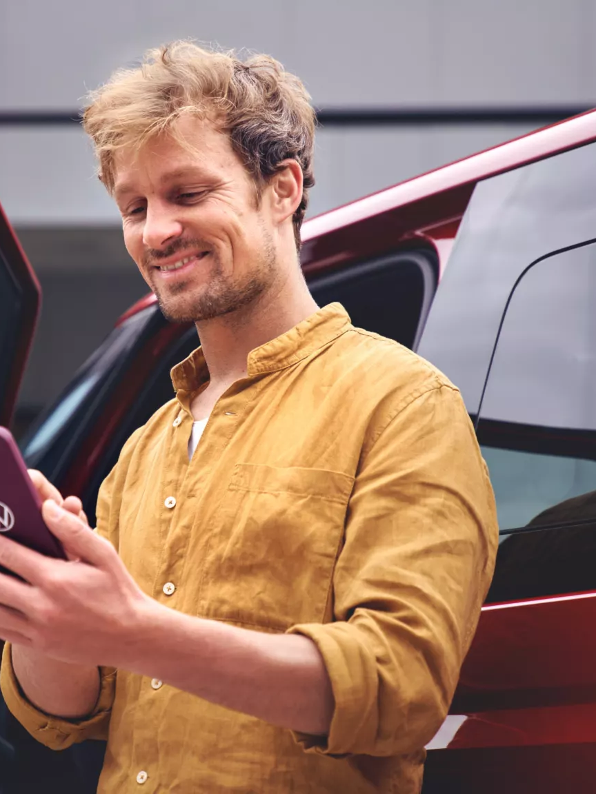 Ein Mann steht neben einem roten Auto und hält ein Smartphone in der Hand. Er trägt ein senfgelbes Hemd.