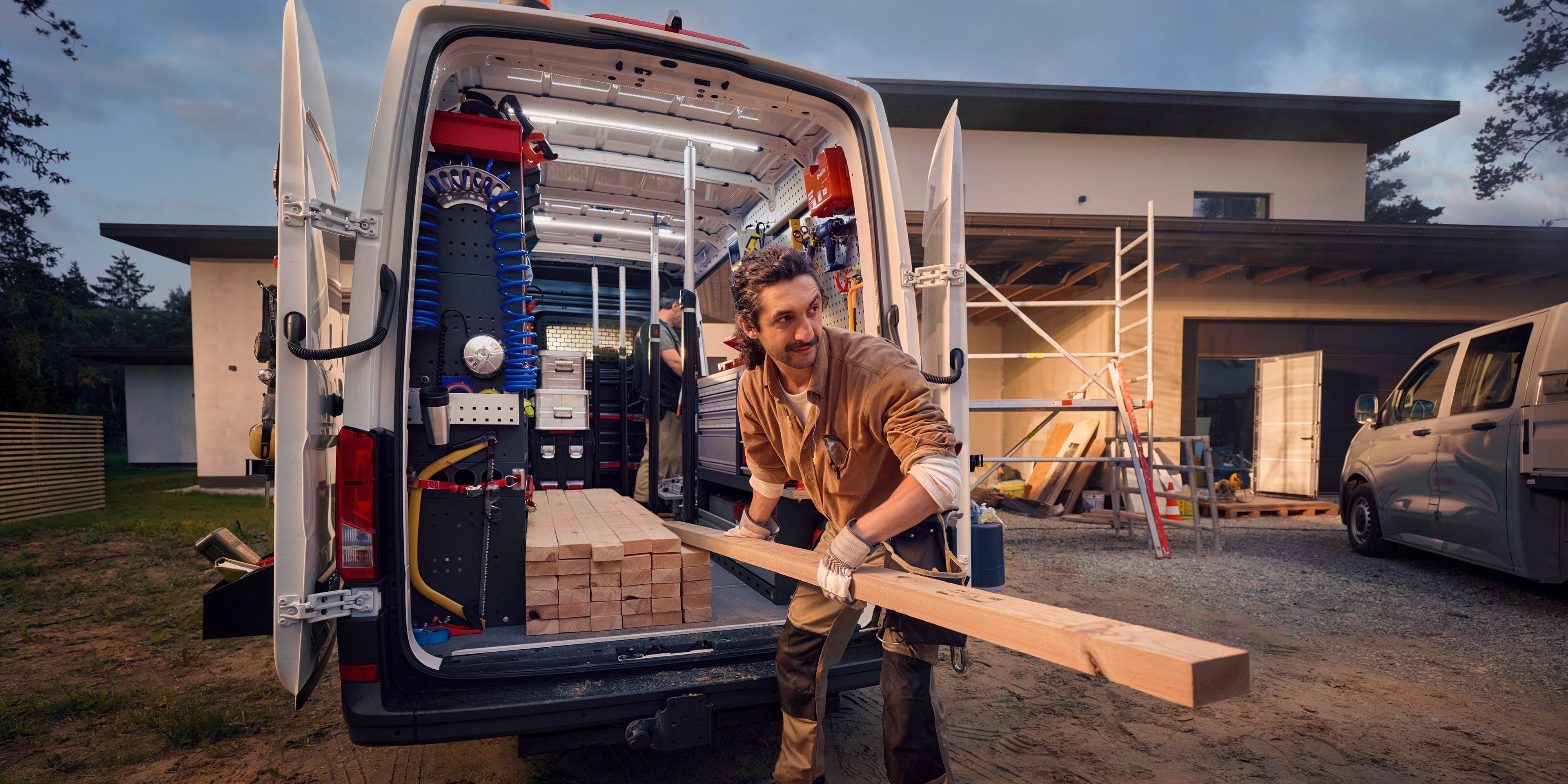 Ein Bauarbeiter lädt Holz aus einem Lieferwagen vor einem Haus, das sich im Bau befindet. Im Hintergrund sind Gerüste und Baumaterialien zu sehen.