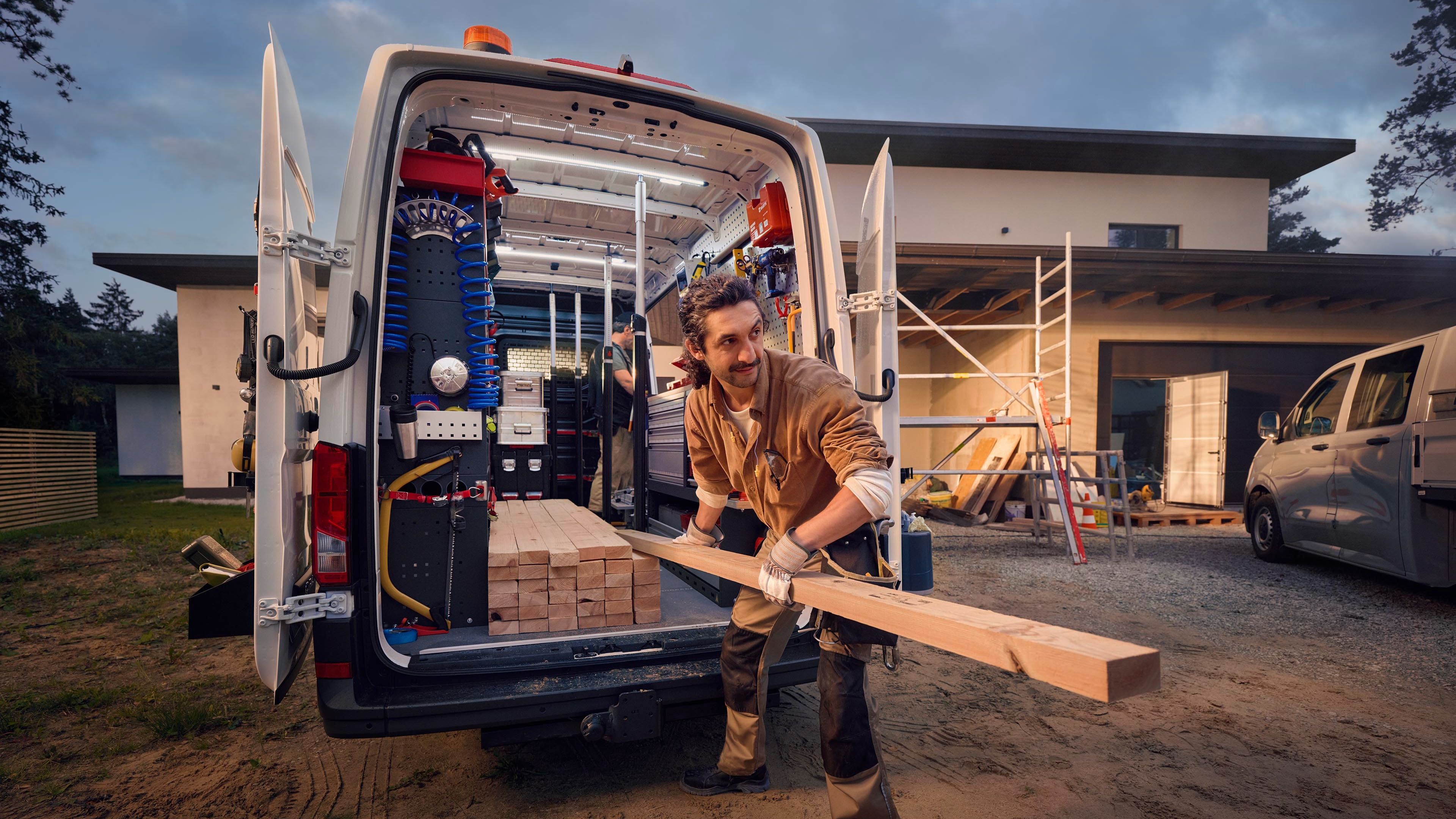 Ein Bauarbeiter lädt Holz aus einem Lieferwagen vor einem Haus, das sich im Bau befindet. Im Hintergrund sind Gerüste und Baumaterialien zu sehen.