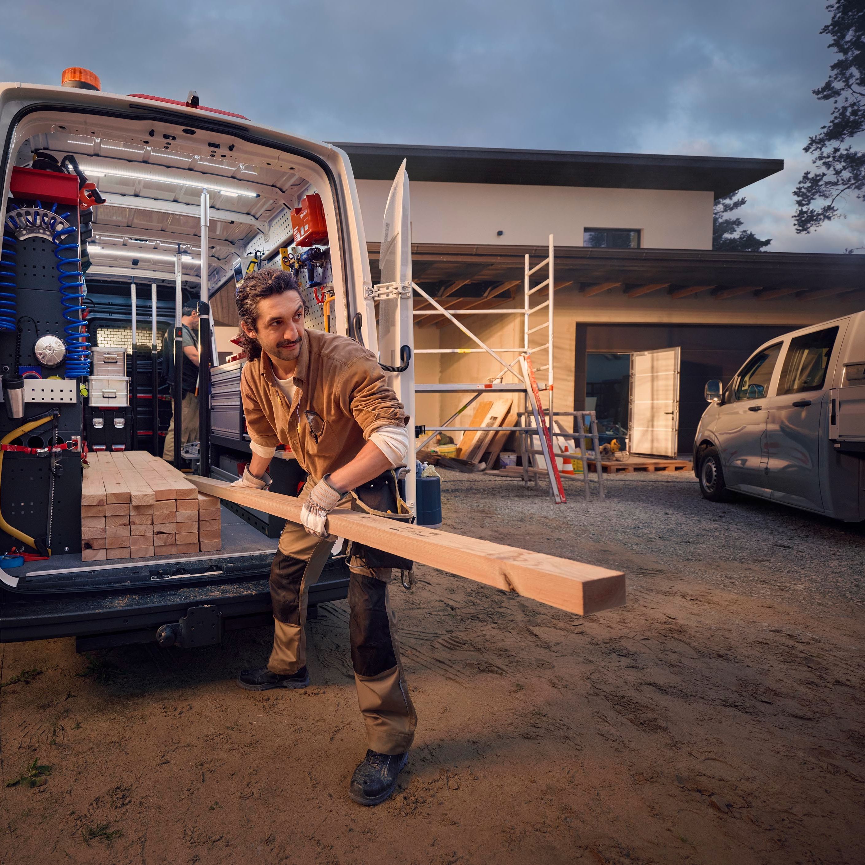 Ein Bauarbeiter lädt Holz aus einem Lieferwagen vor einem Haus, das sich im Bau befindet. Im Hintergrund sind Gerüste und Baumaterialien zu sehen.