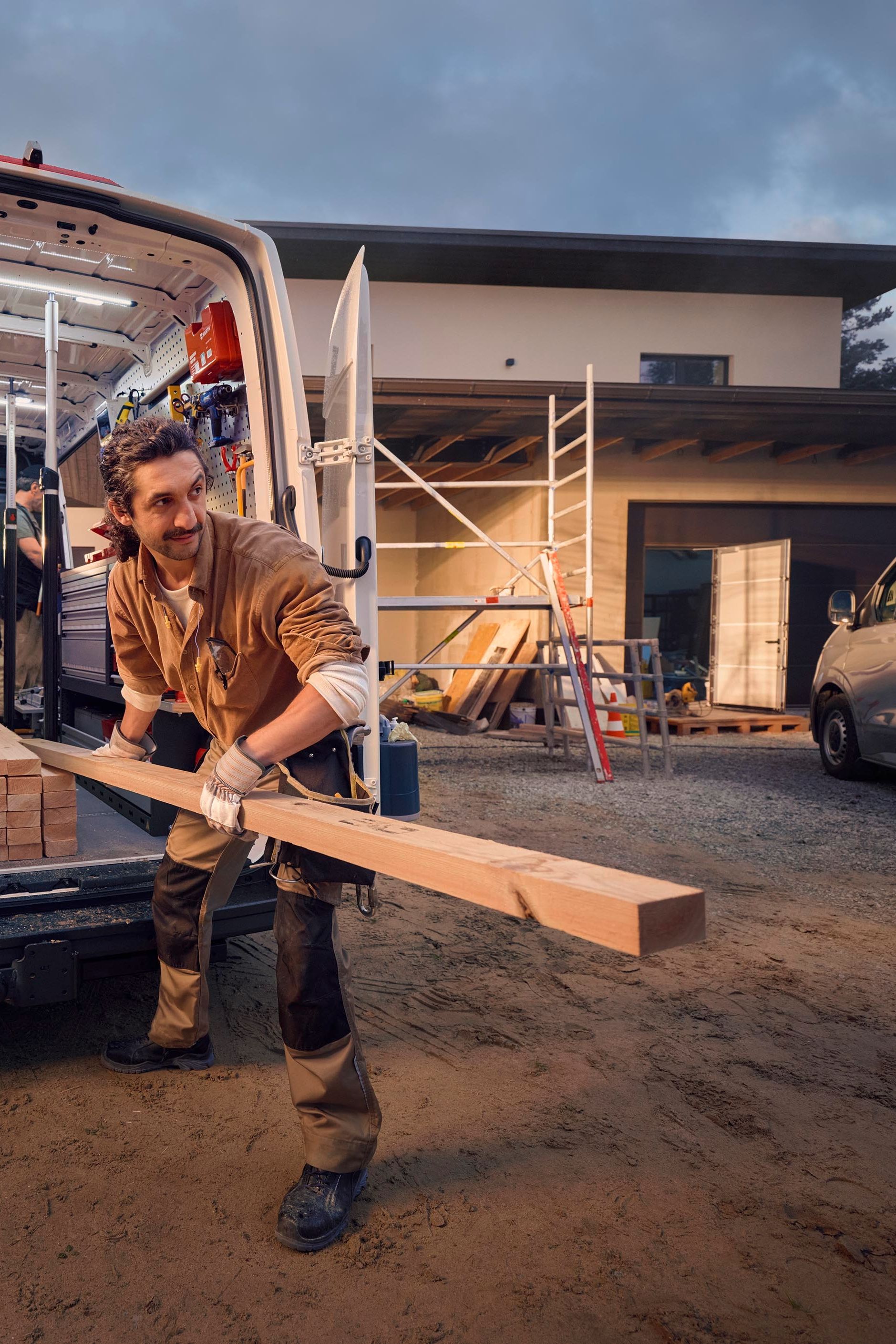 Ein Bauarbeiter lädt Holz aus einem Lieferwagen vor einem Haus, das sich im Bau befindet. Im Hintergrund sind Gerüste und Baumaterialien zu sehen.