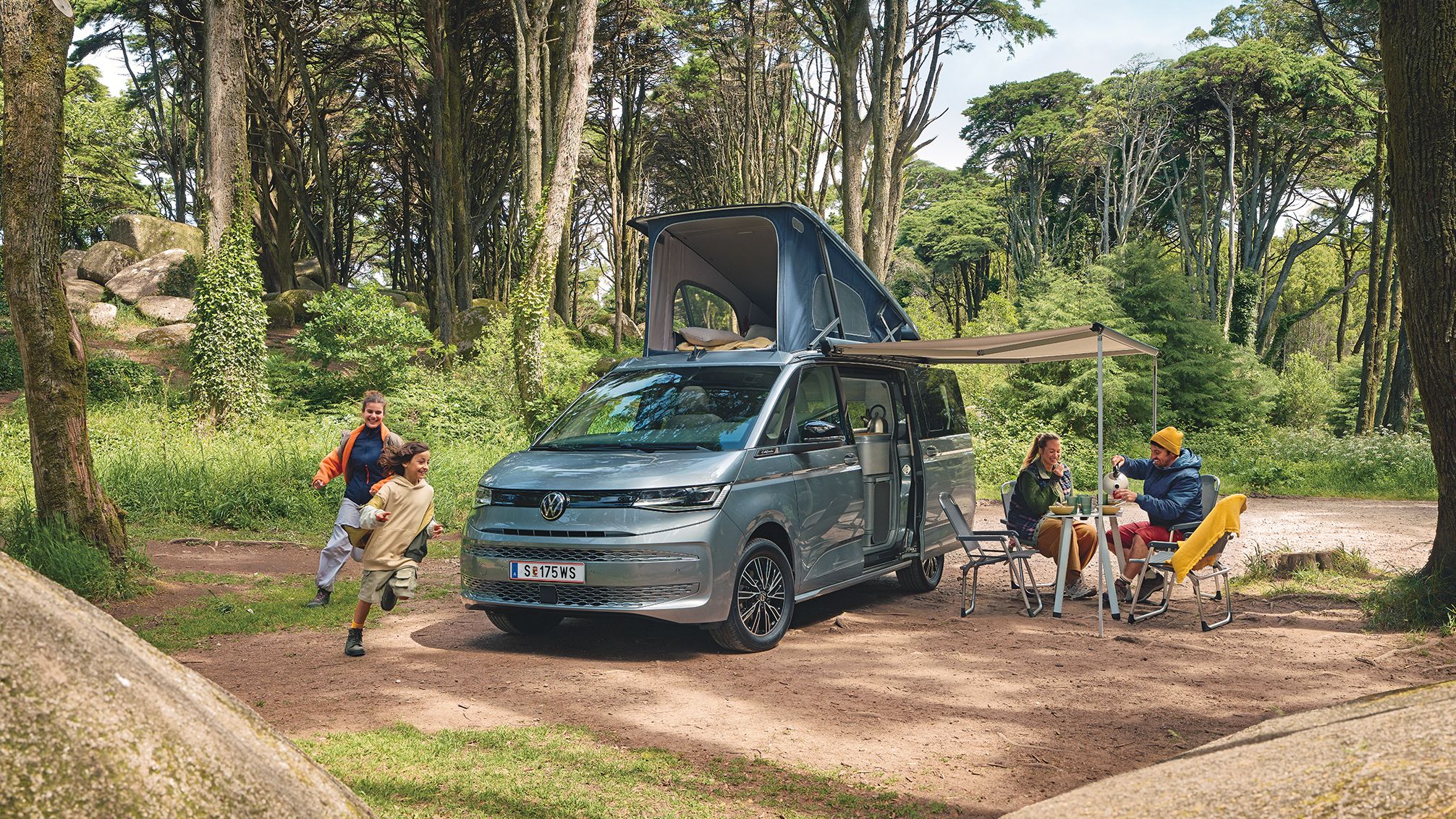 Ein New Multivan parkt im Wald,  zwei Kinder laufen darum herum, die Eltern frühstücken unter der ausgefahrenen Marquise