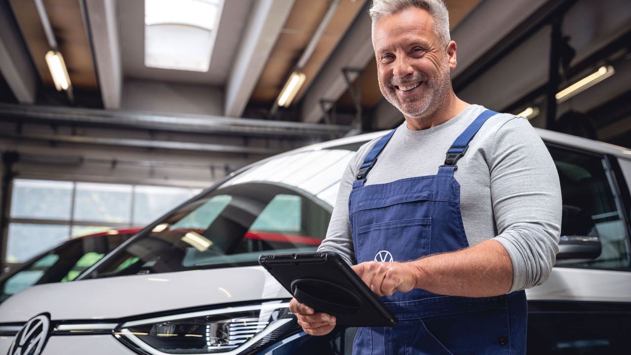 Ein Volkswagen Mechaniker in einer Werkstatt trägt blaue Arbeitskleidung und benutzt ein Tablet. Im Hintergrund steht ein Volkswagen ID. Buzz.
