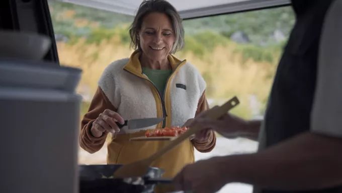 Zwei Personen bereiten Essen in einem Wohnmobil vor. Eine Person hält ein Messer und schneidet Tomaten. Die andere Person hält einen Holzkochlöffel und rührt in einer Pfanne.