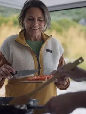 Zwei Personen bereiten Essen in einem Wohnmobil vor. Eine Person hält ein Messer und schneidet Tomaten. Die andere Person hält einen Holzkochlöffel und rührt in einer Pfanne.