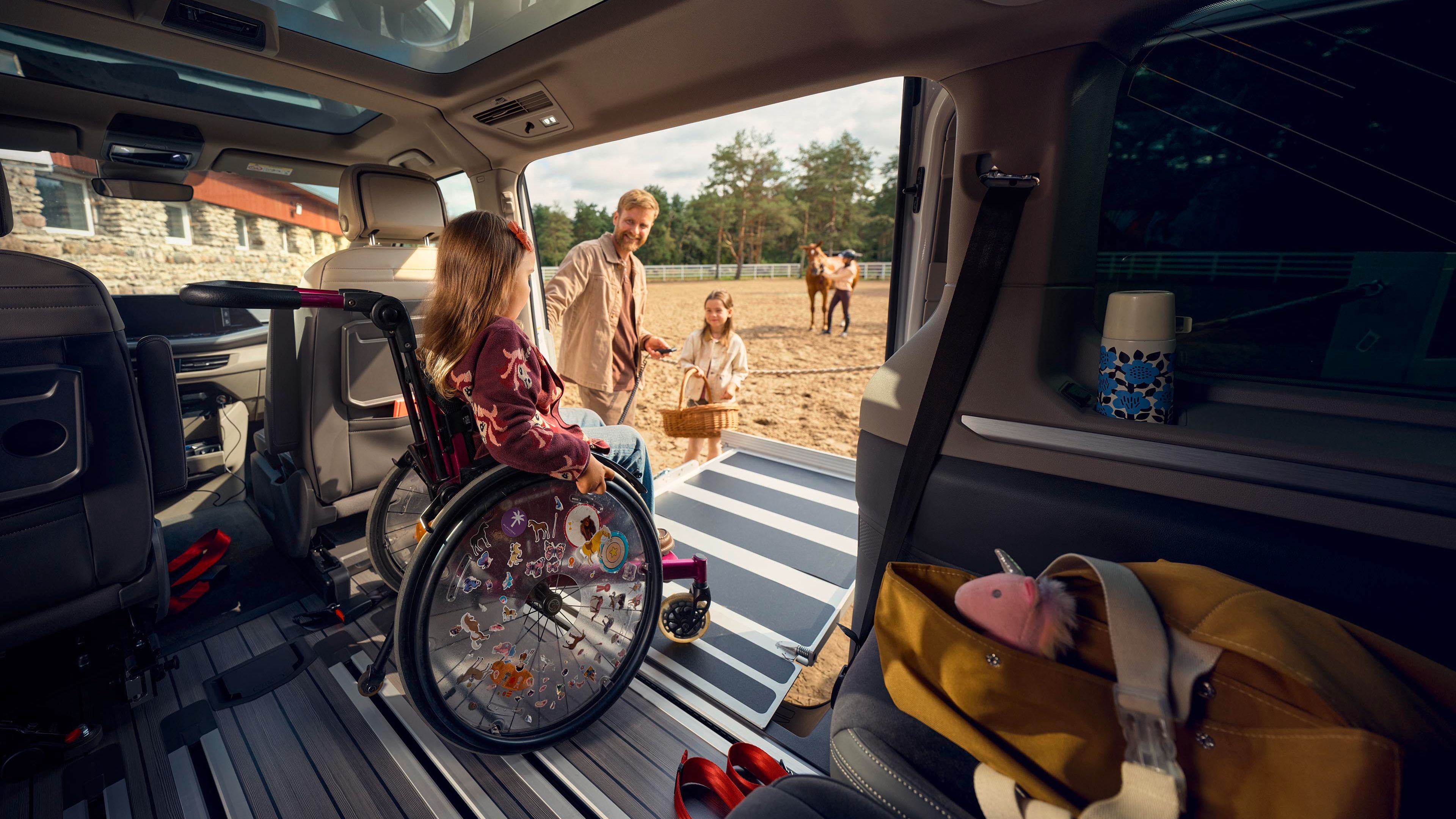 Ein Mädchen im Rollstuhl wird von einem Auto auf einen Sandplatz geschoben. Im Hintergrund sind zwei Erwachsene, die einen Korb tragen, sowie ein Pferd auf einer Koppel zu sehen. Die Innenseite des Autos ist teilmöbliert, einschließlich einer Tasche mit verschiedenen Gegenständen.