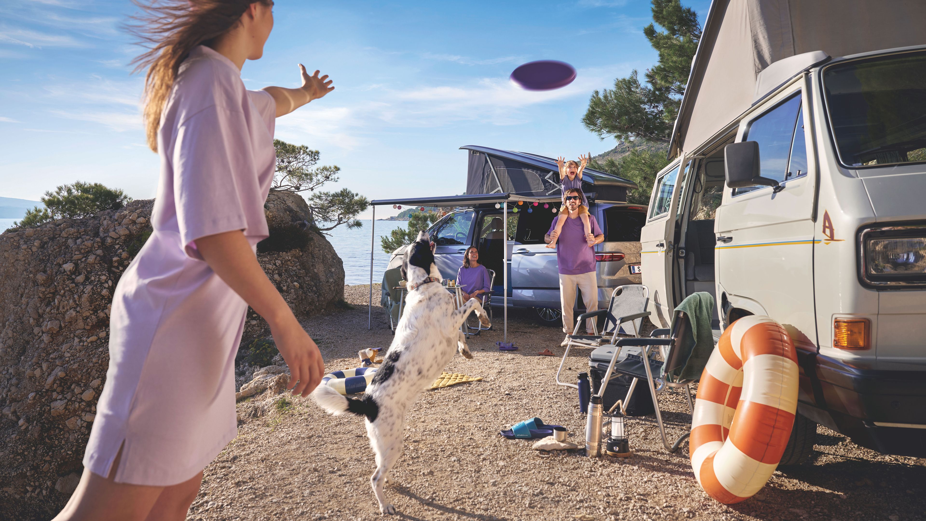 Eine Familie spielt mit ihrem Hund auf einem Campingplatz an der Küste. Im Hintergrund sieht man zwei Volkswagen California.