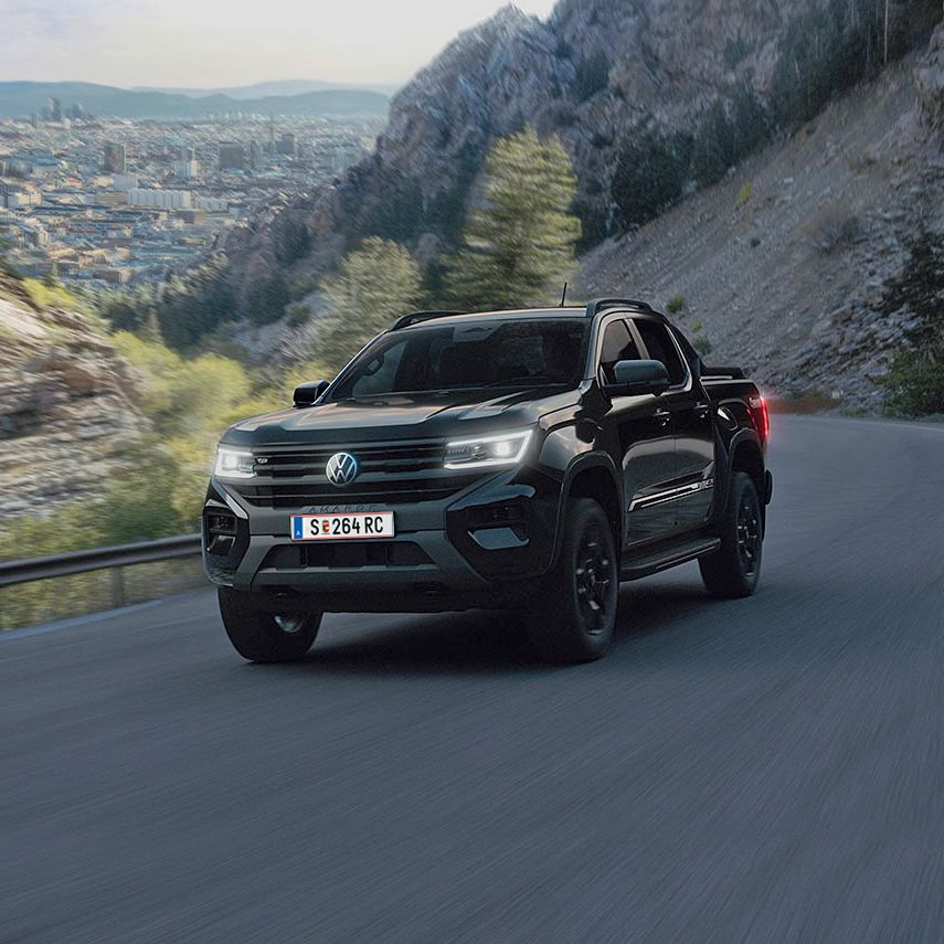 Ein schwarzer Pickup fährt entlang einer kurvenreichen Bergstraße. Im Hintergrund sind steile Felsen und eine Landschaft mit Bäumen und einer Stadt zu sehen. Der Himmel ist teilweise bewölkt.