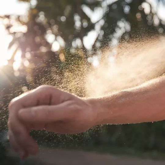 Ein Mensch sprüht Insektenschutzmittel auf seinen ausgestreckten Arm. Der Hintergrund zeigt unscharfe Bäume und Sonnenschein.