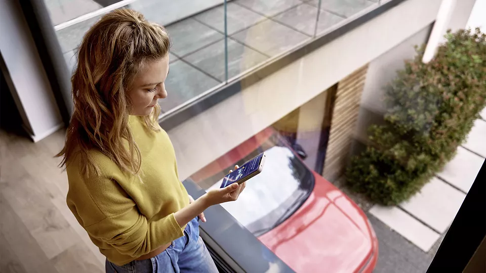 Junge Frau mit blondem, halblangem Haar steht in einem modernen Haus an einer großen Glasscheibe und schaut auf ein Smartphone. Draußen unten ist ein rotes Auto in einer Einfahrt oder Garage zu sehen. Die Szene wirkt hell und modern, mit minimalistischer Einrichtung und Glasbalkon.