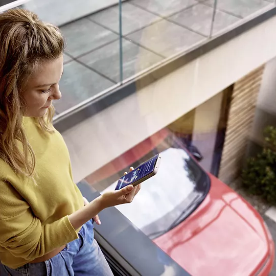 Junge Frau mit blondem, halblangem Haar steht in einem modernen Haus an einer großen Glasscheibe und schaut auf ein Smartphone. Draußen unten ist ein rotes Auto in einer Einfahrt oder Garage zu sehen. Die Szene wirkt hell und modern, mit minimalistischer Einrichtung und Glasbalkon.