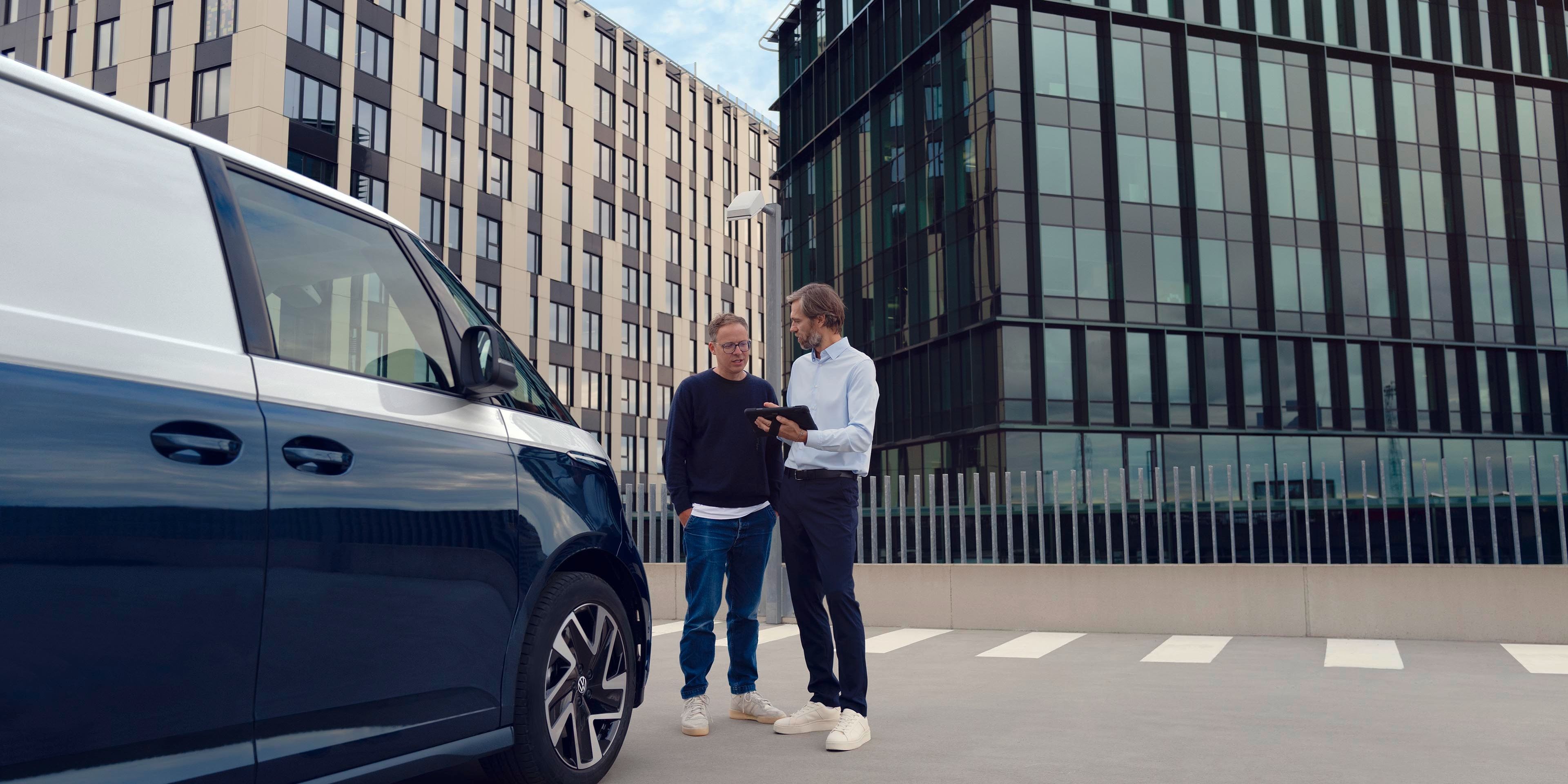 Zwei Personen stehen neben einem blauen Lieferwagen auf einem Parkplatz, umgeben von modernen Bürogebäuden. Eine Person zeigt der anderen etwas auf einem Tablet.