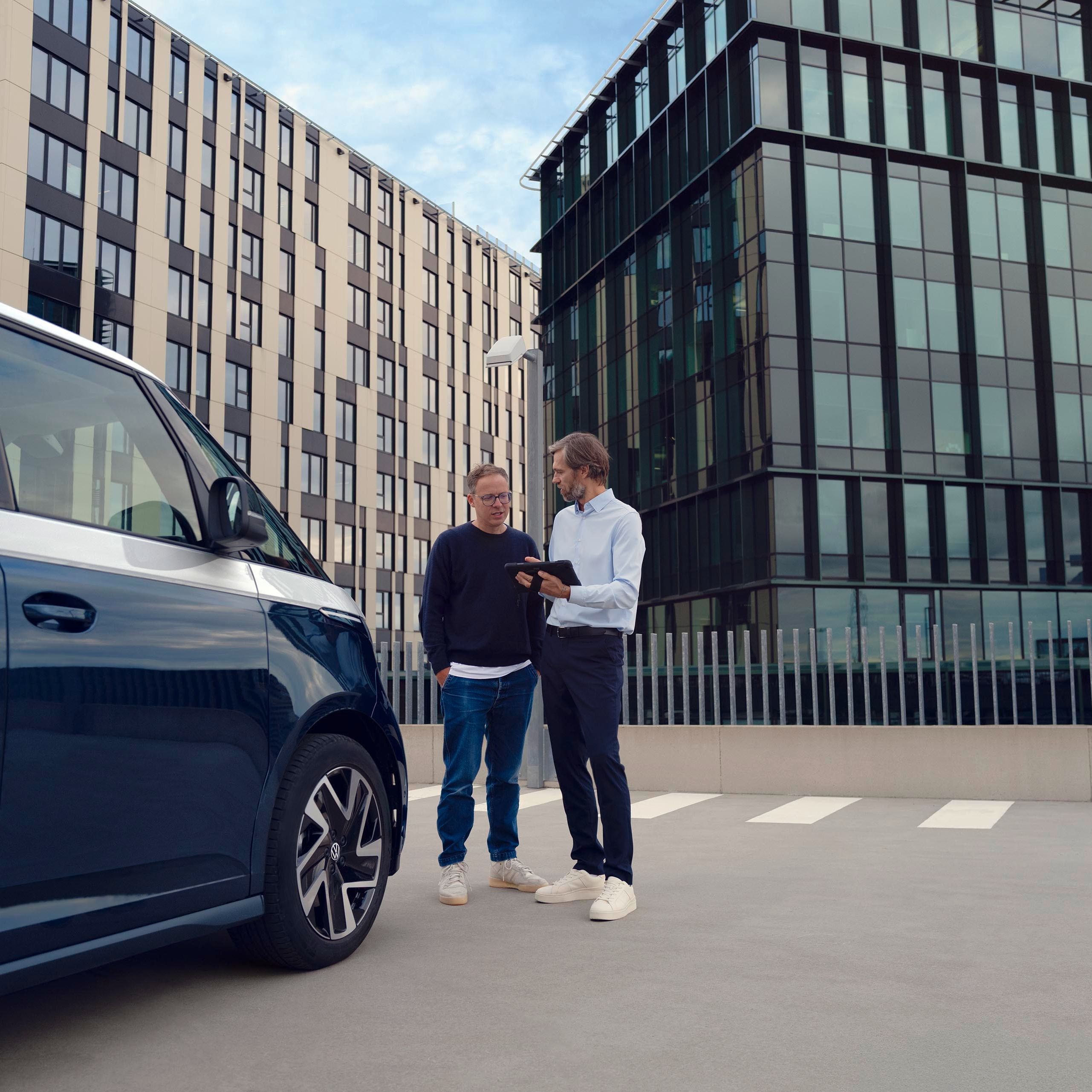 Zwei Personen stehen neben einem blauen Lieferwagen auf einem Parkplatz, umgeben von modernen Bürogebäuden. Eine Person zeigt der anderen etwas auf einem Tablet.
