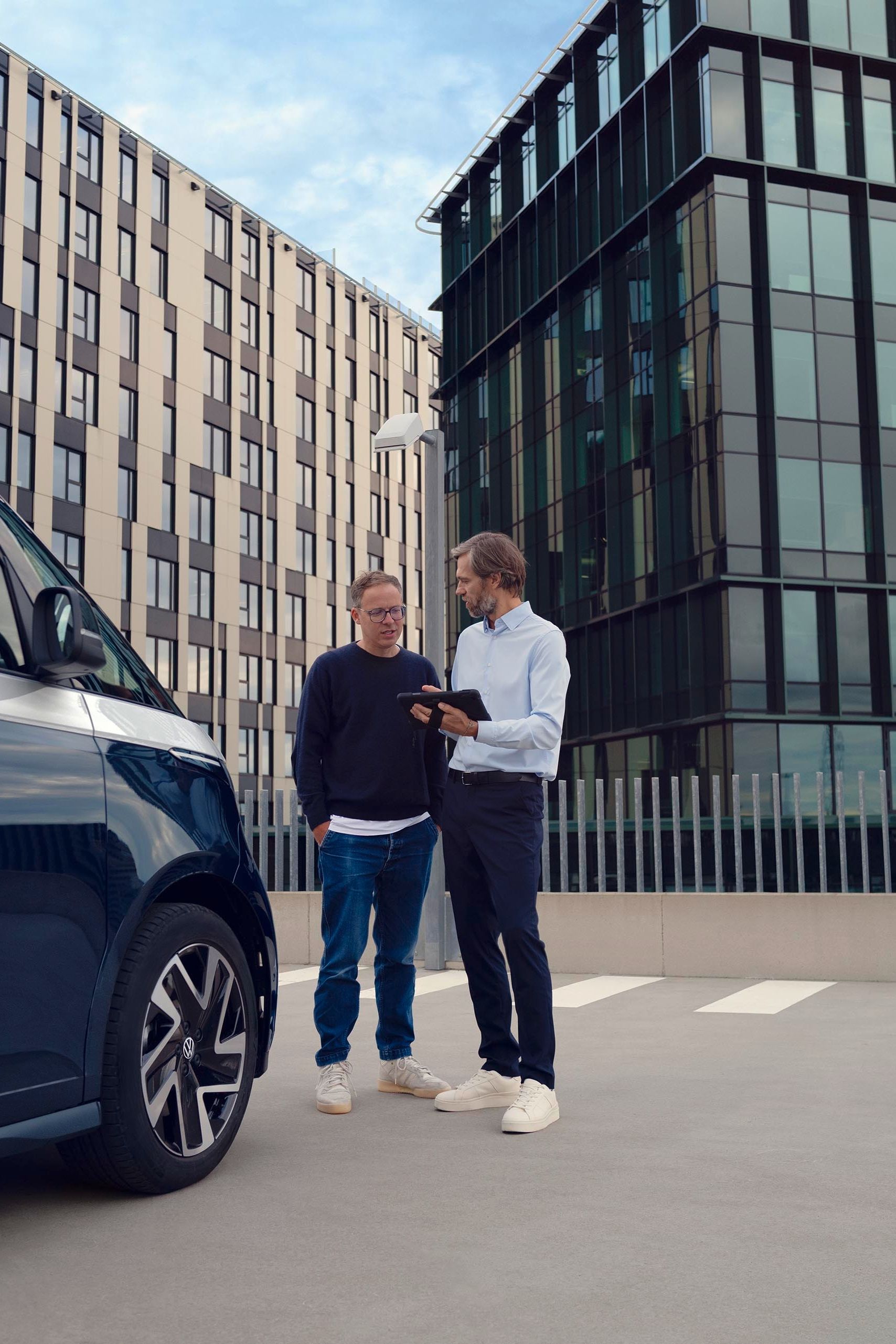 Zwei Personen stehen neben einem blauen Lieferwagen auf einem Parkplatz, umgeben von modernen Bürogebäuden. Eine Person zeigt der anderen etwas auf einem Tablet.