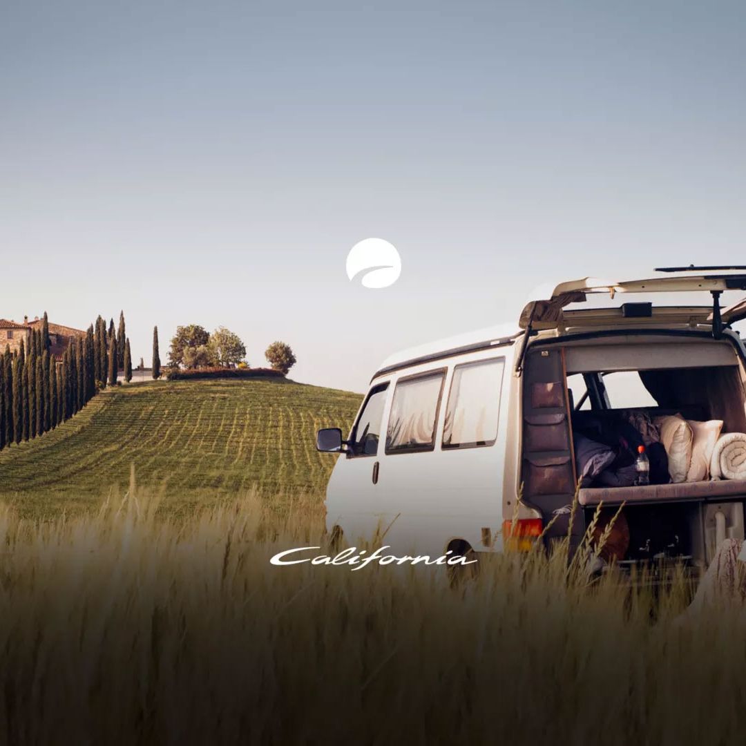 Ein VW Transporter ist auf einem Feld geparkt mit Blick auf eine Reihe von Zypressen und ein Haus auf einem Hügel. Eine Person sitzt neben dem VW und genießt die Landschaft. Im Hintergrund ist der Himmel klar und blau.