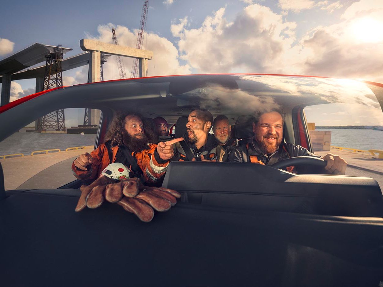 Mehrere Personen sitzen in einem Auto und tragen Sicherheitsanzüge. Im Vordergrund ist ein Kinderspielzeug in Form einer Krabbe zu sehen. Im Hintergrund sind Baustellenstrukturen und ein Wassergebiet ersichtlich.