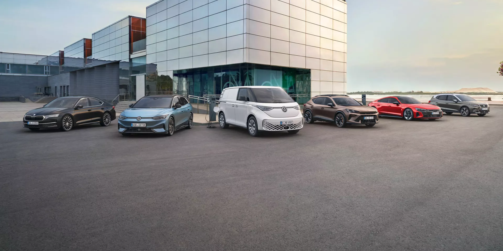 Eine Reihe von Fahrzeugen steht vor einem modernen Gebäude. Es sind verschiedene Autotypen, darunter Limousinen, ein Van und ein Sportwagen. Die Autos sind in unterschiedlichen Farben lackiert.