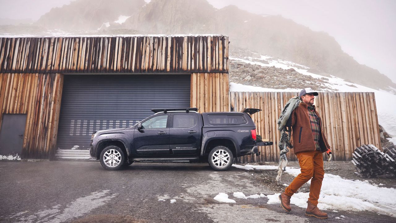 Ein Mann in Outdoor-Kleidung und mit einem großen Rucksack geht auf einer nassen, teils verschneiten Straße an einem schwarzen Volkswagen Amarok vorbei. Der Amarok steht vor einem modernen Holzgebäude mit großem Rolltor.